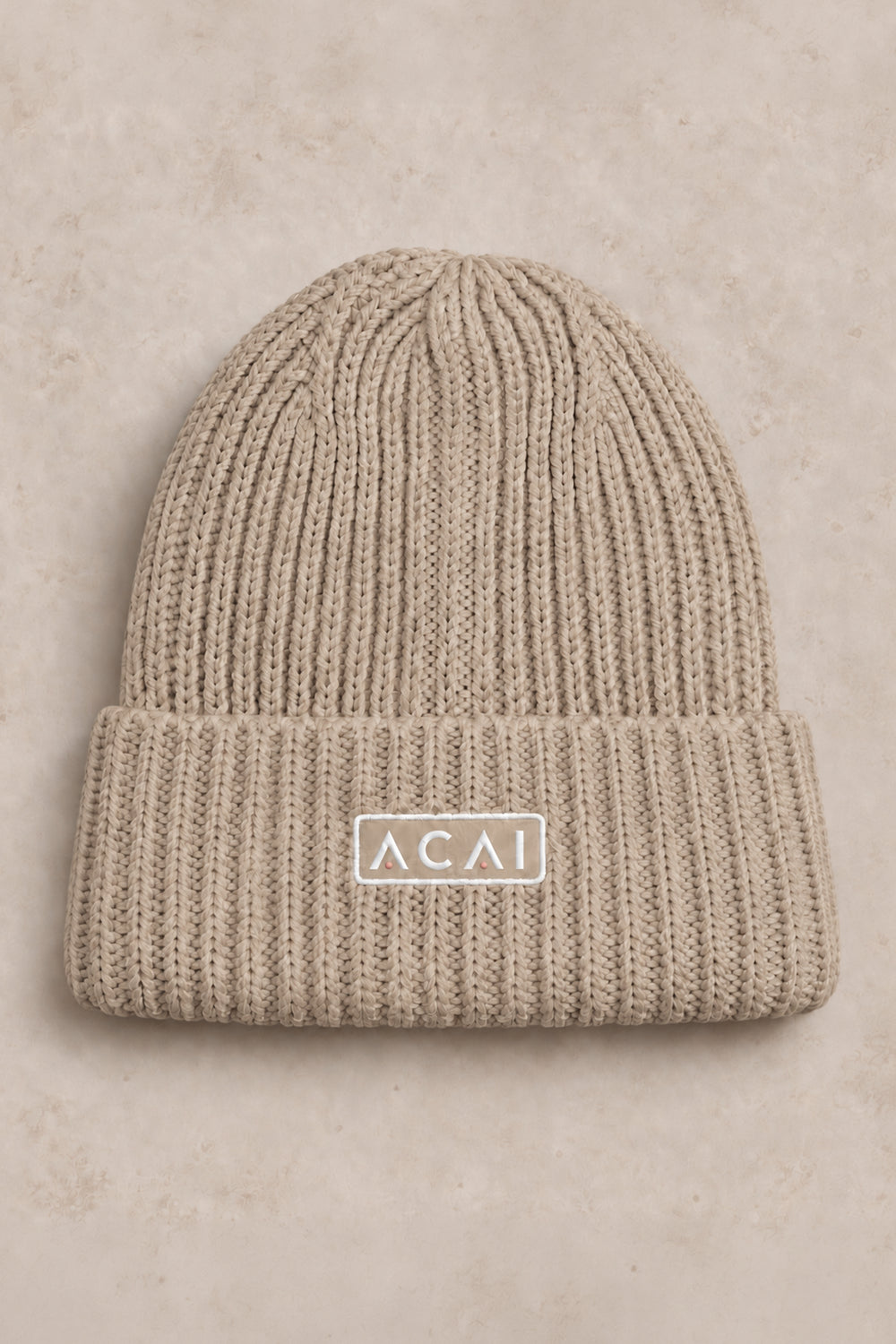 A beige ribbed knit beanie hat with a folded cuff and a small white rectangular patch reading 'ACAI' in the center, shown on a textured beige background.