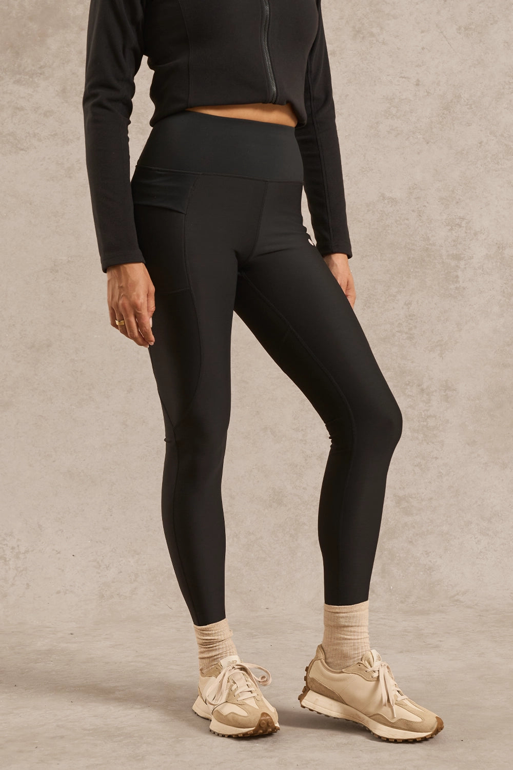 A model wearing black high-waisted athletic leggings, a black cropped long-sleeve top, beige socks, and beige sneakers, standing against a textured light gray studio backdrop.