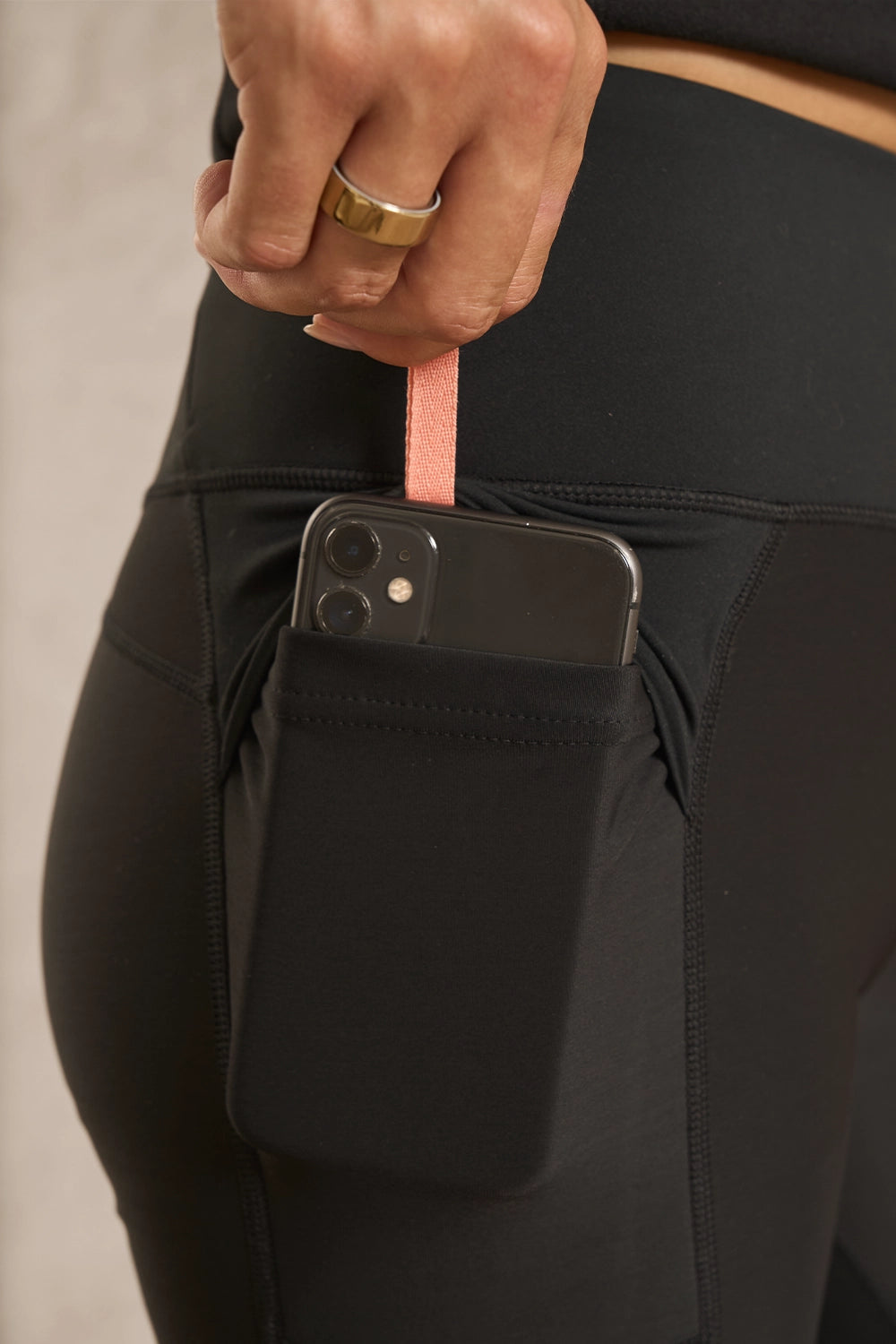 Close-up of a person wearing black athletic leggings with a side pocket holding a smartphone, with a pink wrist strap hanging from the pocket and a hand reaching in.