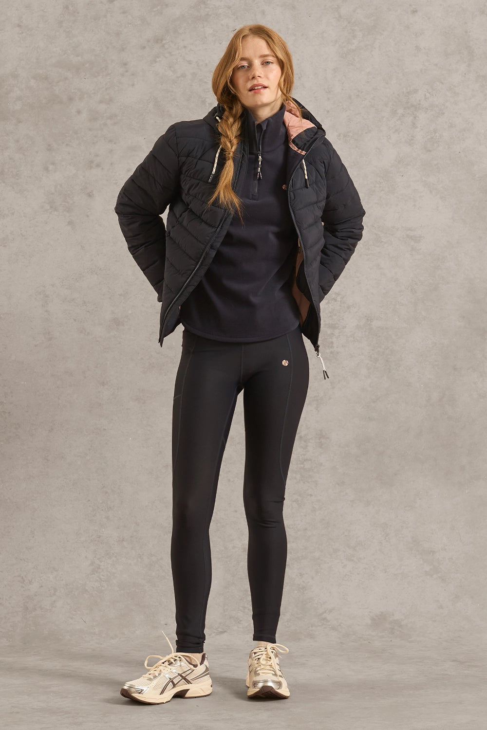 A woman modeling a black puffer jacket over a black top and black leggings, standing against a textured gray studio backdrop.