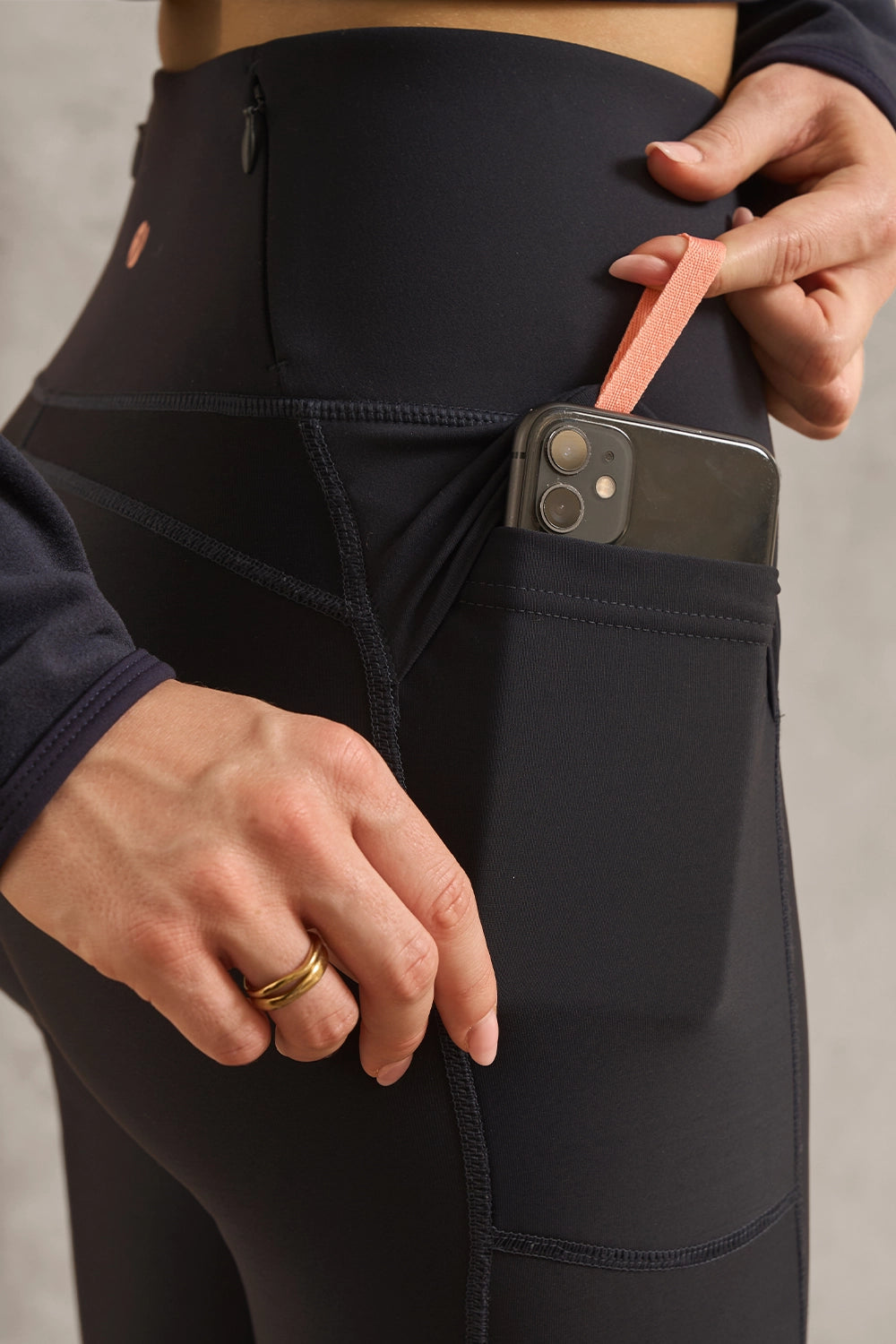 Close-up of a person wearing black high-waisted athletic leggings with a side pocket holding a smartphone and a peach wrist strap, against a neutral gray background.