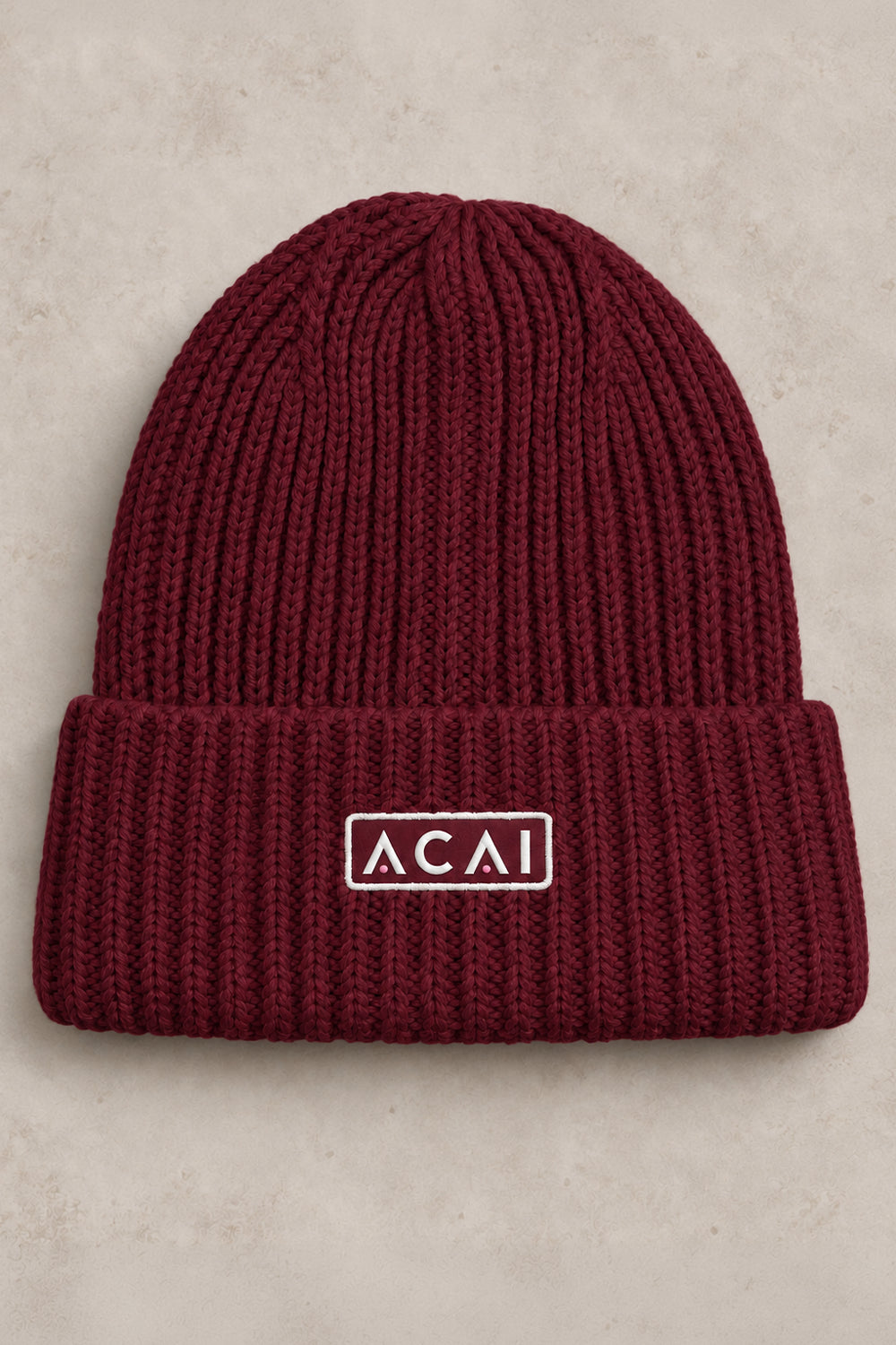 A ribbed knit cuffed beanie in deep burgundy with a white rectangular logo patch reading 'ACAI' on the front cuff, photographed flat against a light beige textured background.