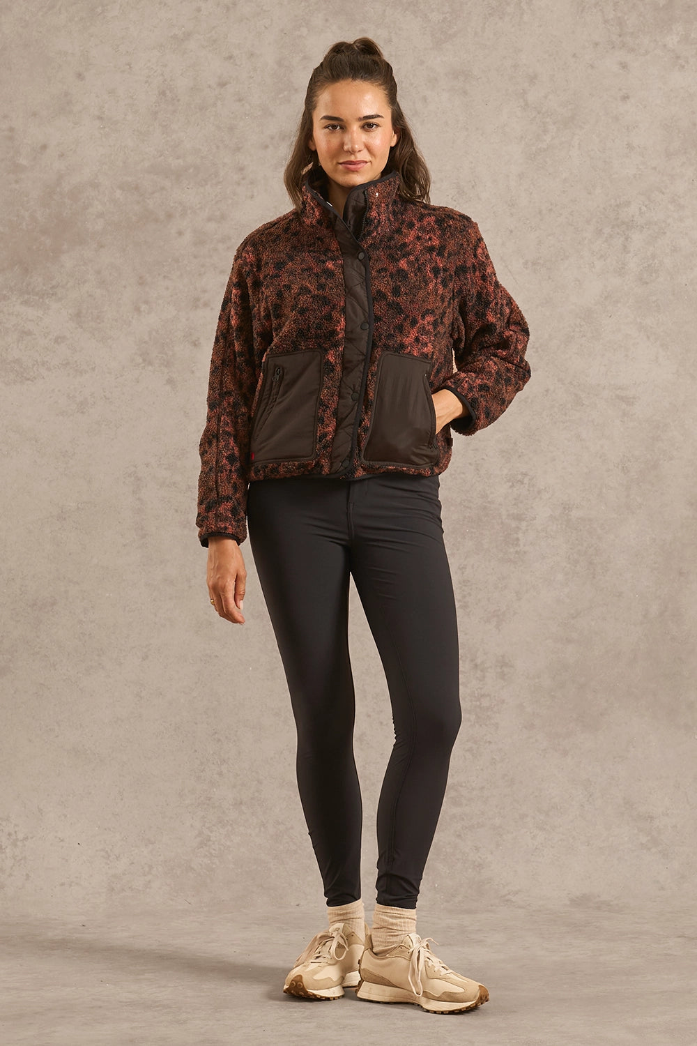 A woman modeling a cropped zip-up jacket with a dark brown and black abstract pattern, paired with black leggings and beige sneakers, standing against a textured gray studio backdrop.