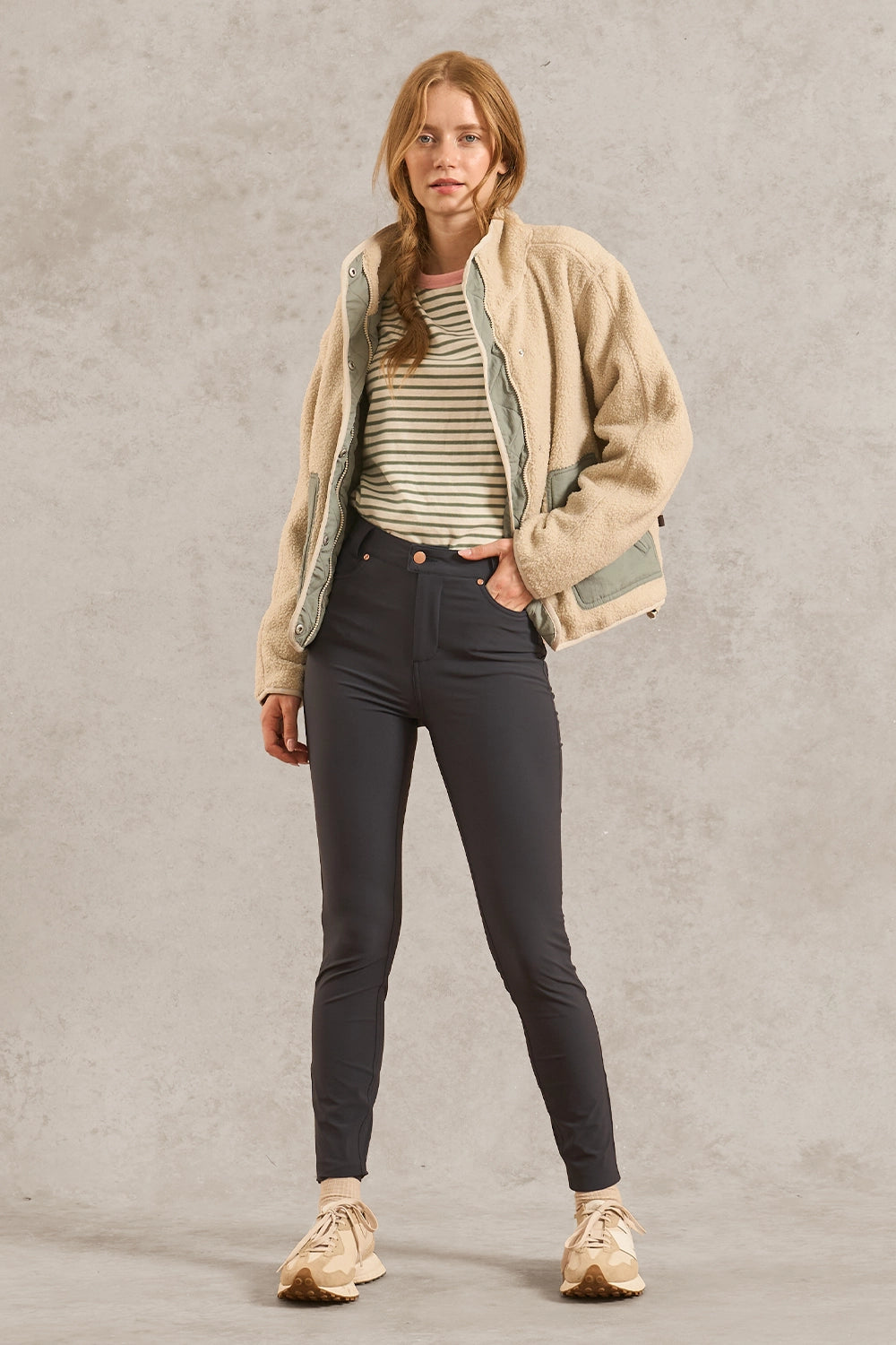 A woman stands against a textured gray studio backdrop wearing a beige sherpa jacket over a striped top, dark gray skinny pants, and beige sneakers.