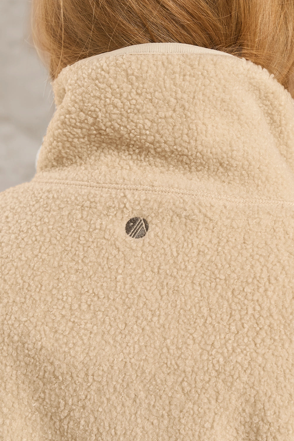 Close-up of the back of a beige sherpa fleece jacket with a small circular logo patch on the upper back, worn by a model with light brown hair.