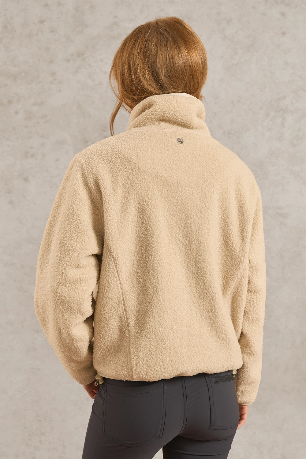 A woman viewed from behind wearing a beige sherpa fleece pullover jacket and dark gray pants, standing against a textured light gray wall.