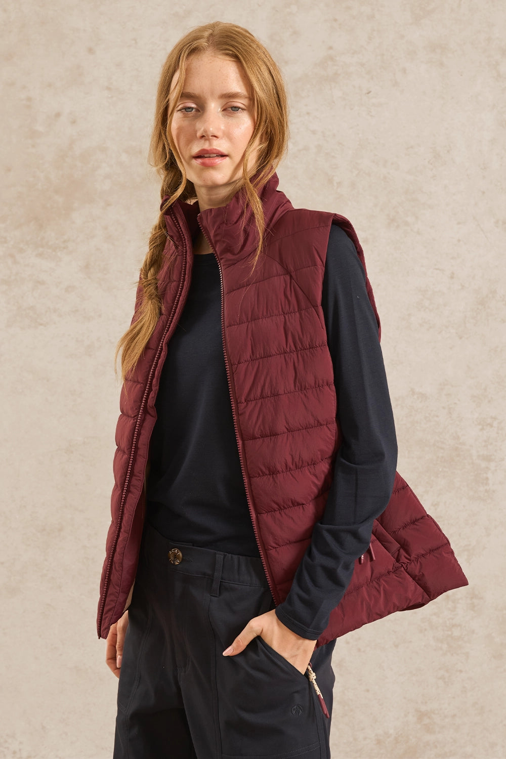 A woman wearing a sleeveless quilted puffer vest in burgundy over a black long-sleeve top and dark pants, standing against a textured beige studio background.