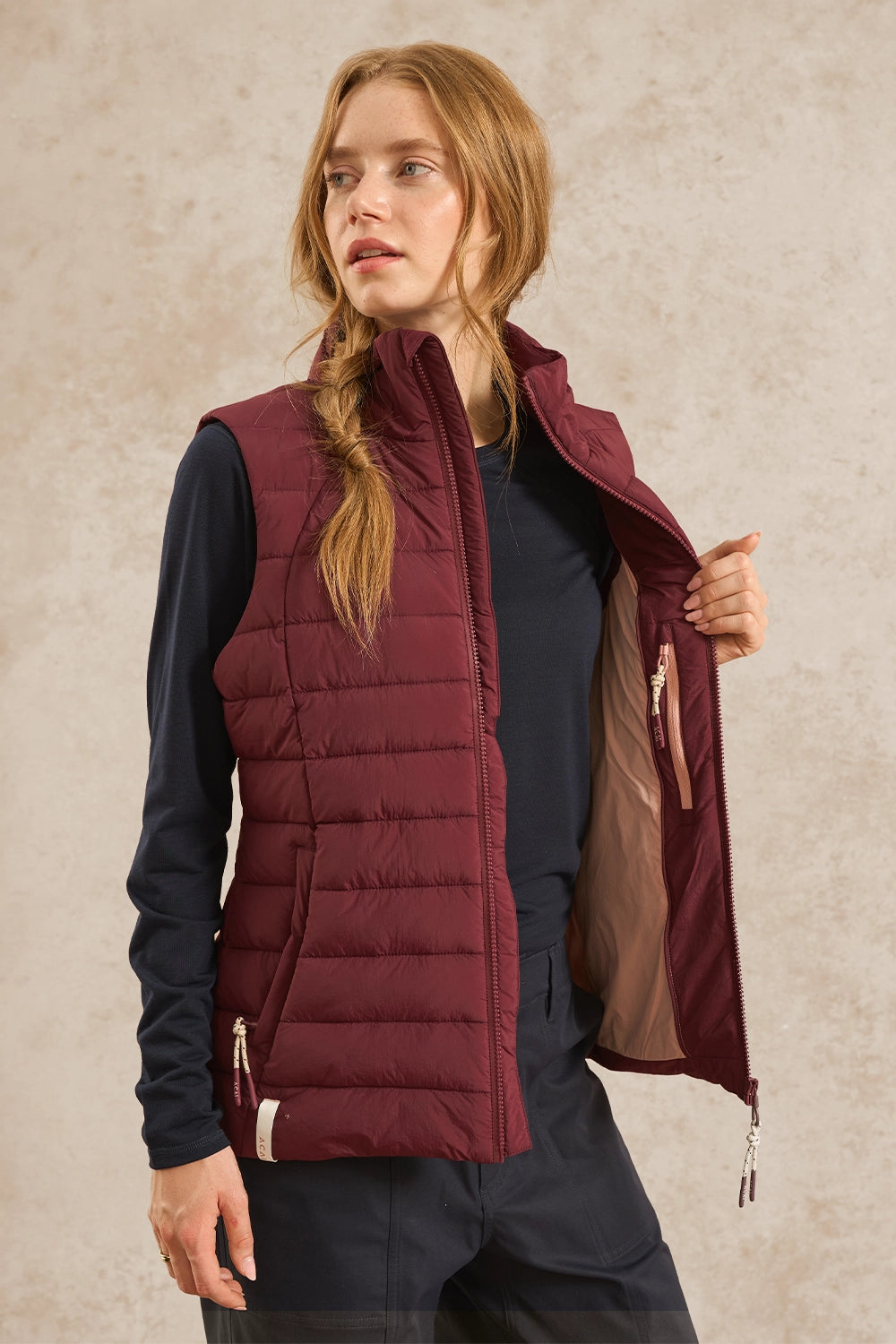 A woman wearing a sleeveless quilted puffer vest in burgundy over a dark navy long-sleeve shirt and navy pants, holding open the vest to show the tan lining. She is standing against a textured beige wall.