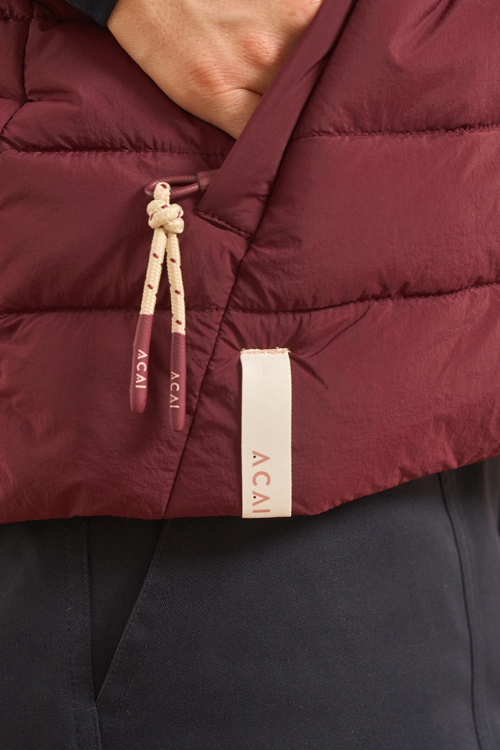 Close-up of a burgundy puffer vest with a cream drawstring and a cream fabric tag printed with the brand name 'ACAI'.
