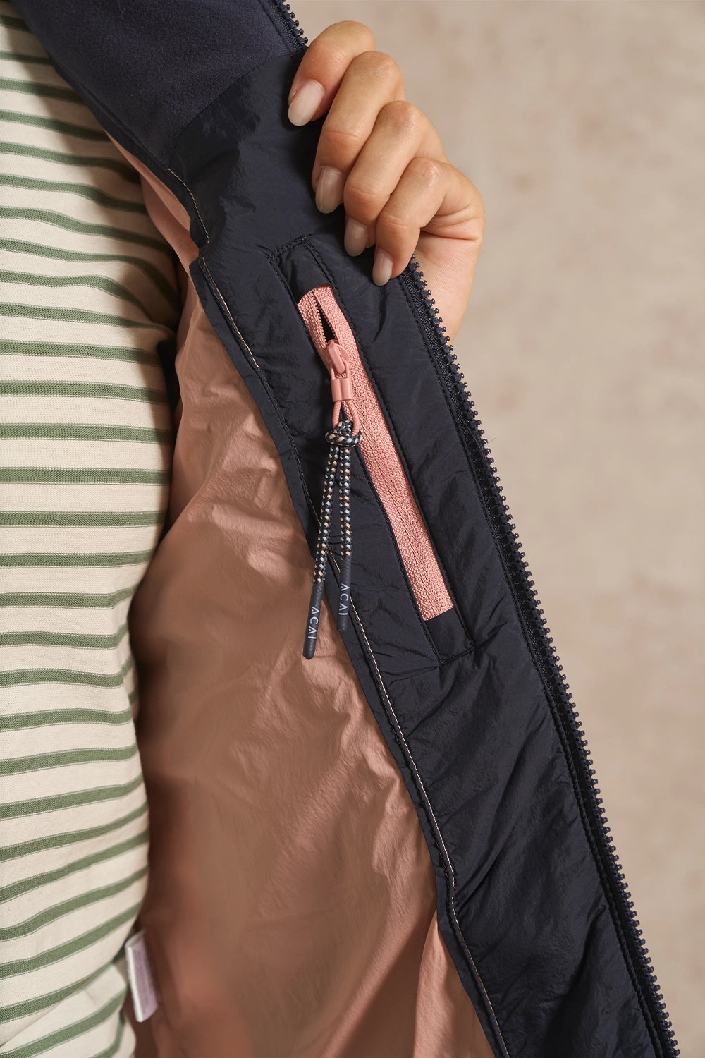 Close-up of a person holding open a dark navy puffer jacket to show the dusty pink inner lining and a pink zipper pull, worn over a green and white striped shirt.