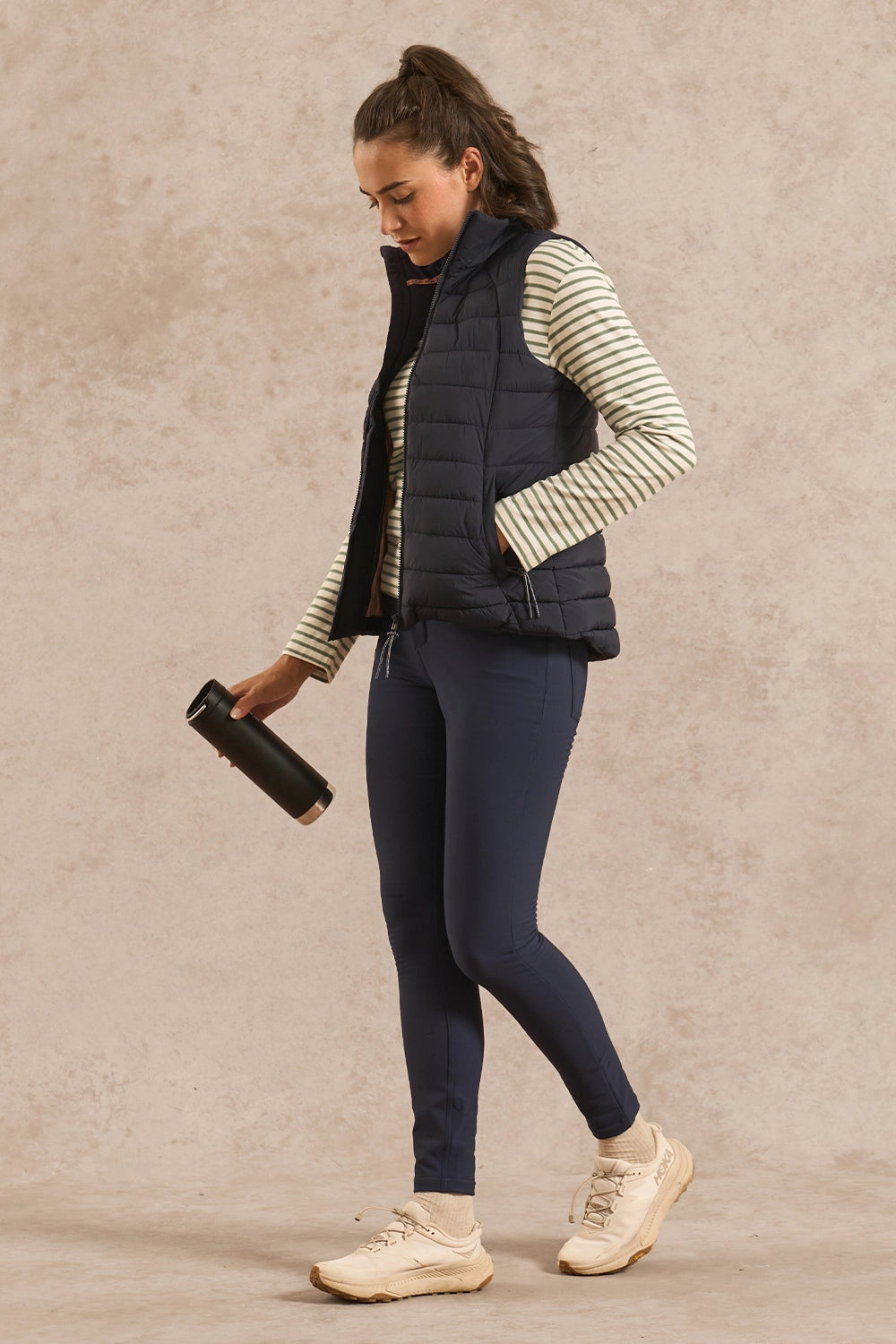 A woman modeling a dark navy puffer vest over a cream and navy striped long-sleeve shirt, paired with navy leggings and beige sneakers, holding a black tumbler against a textured beige studio backdrop.