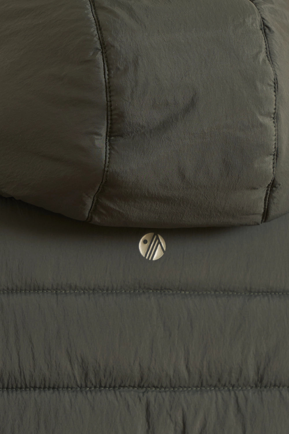 Close-up of a dark olive green puffer jacket showing the quilted fabric texture and a small circular metal logo badge.