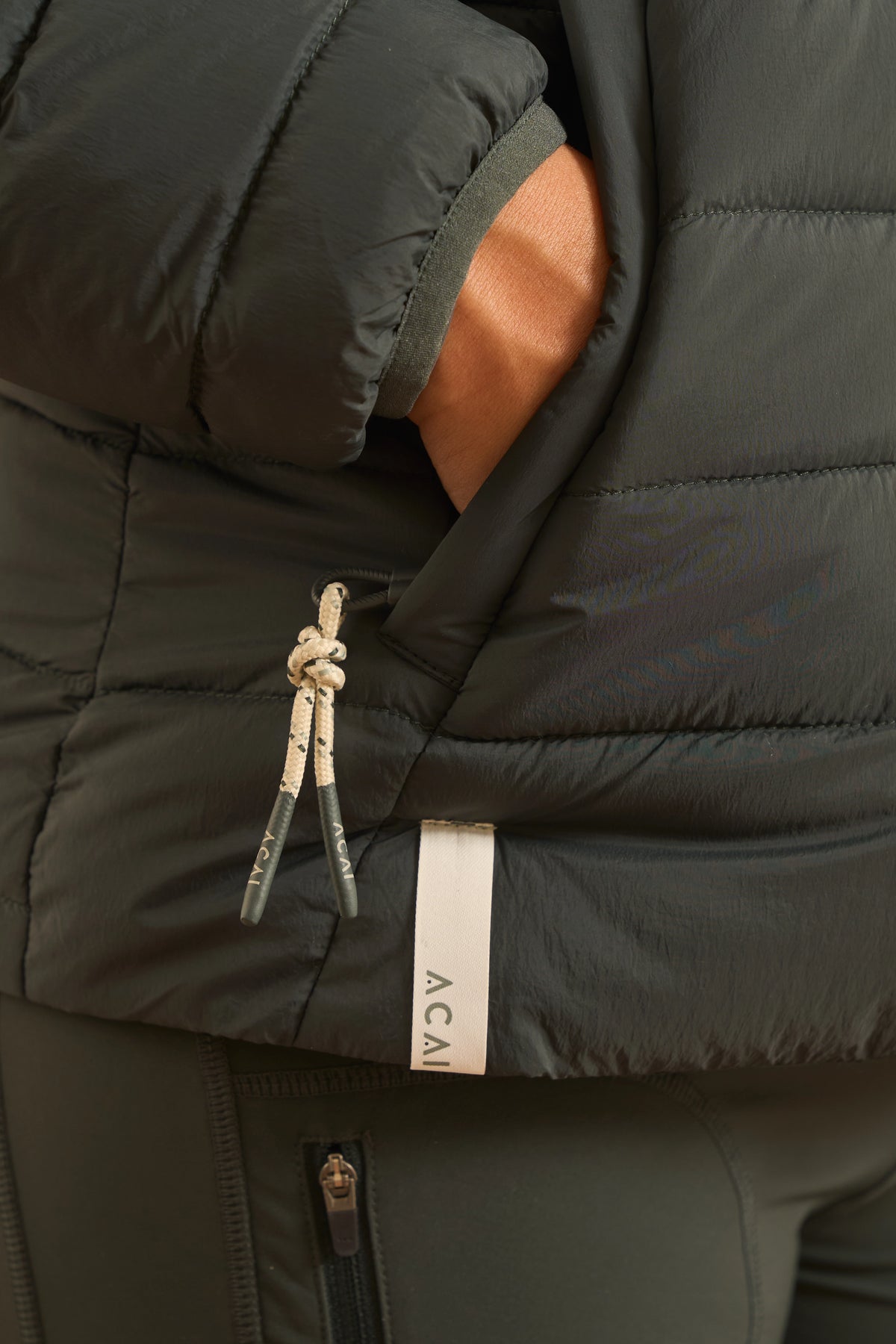Close-up of a dark olive green puffer jacket with a white and gray patterned drawstring and a white vertical brand tag reading ACAI.