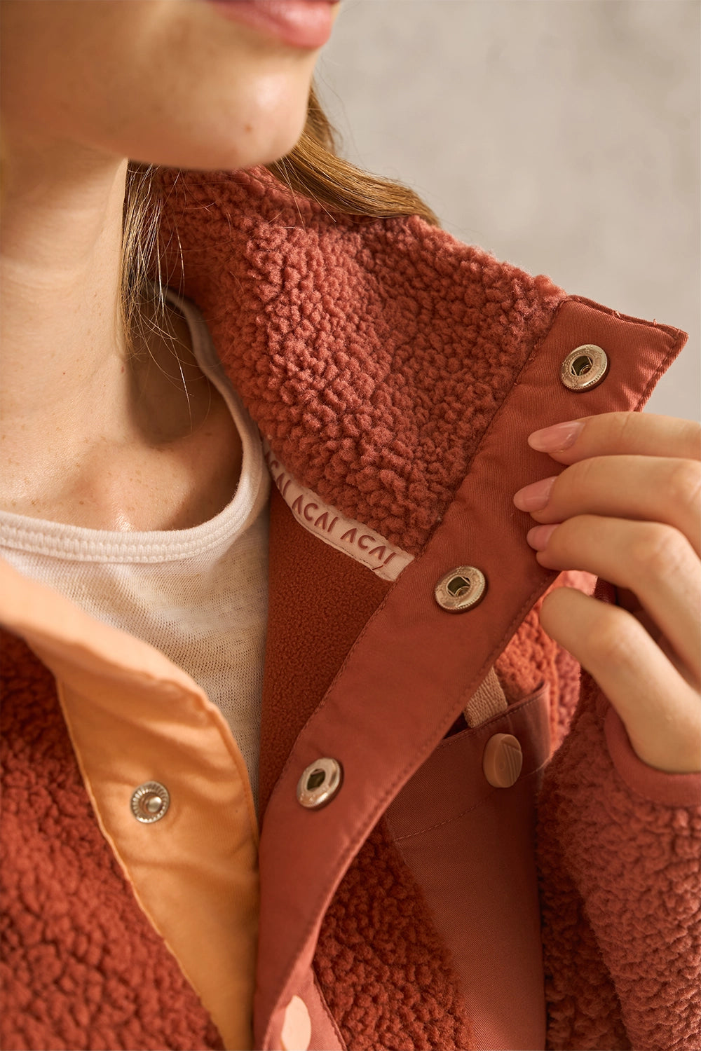 Close-up of a person wearing a rust-colored sherpa fleece jacket with a tan inner layer and metal snap buttons, with a hand adjusting the collar.