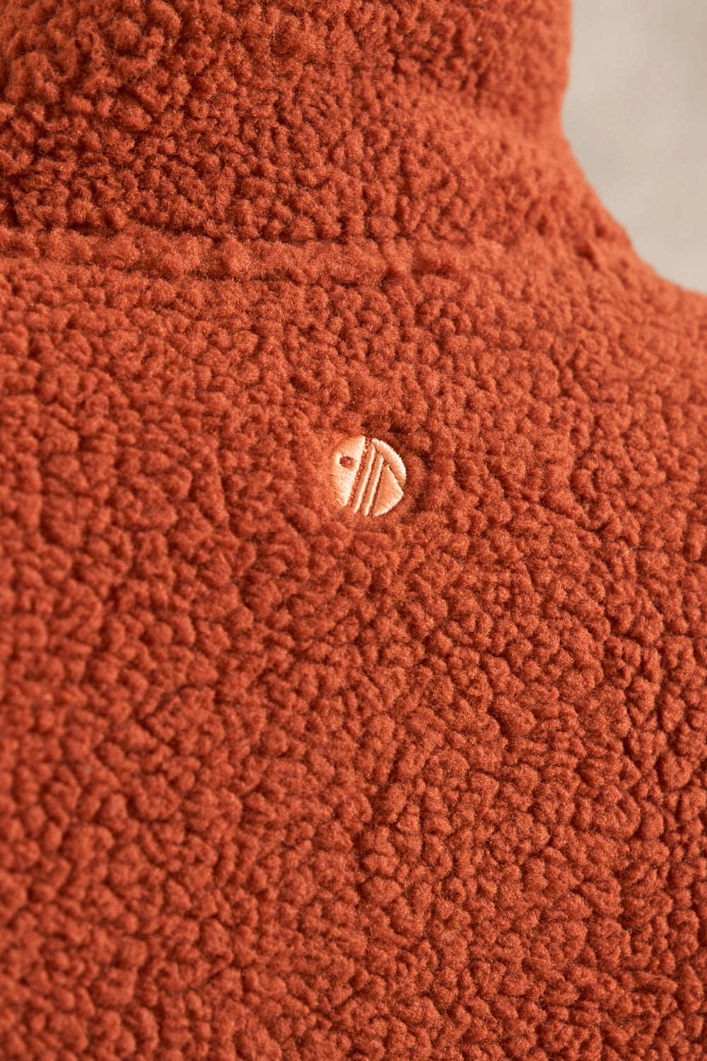Close-up of a textured terracotta fleece garment with a small circular metallic logo patch near the center.