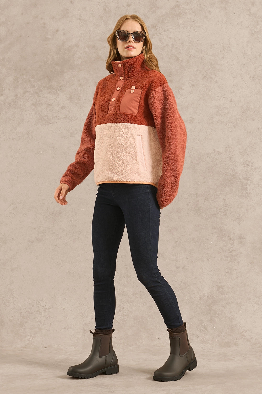 A woman modeling a colorblock fleece pullover sweater with a rust orange top half, a pale pink lower half, and matching chest pocket details, paired with dark skinny jeans and dark brown boots against a textured gray studio backdrop.