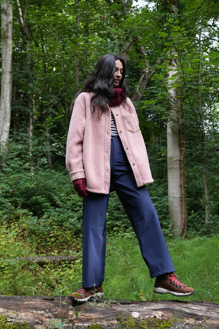 Person standing on a log in a forest wearing a pink coat and blue jeans.