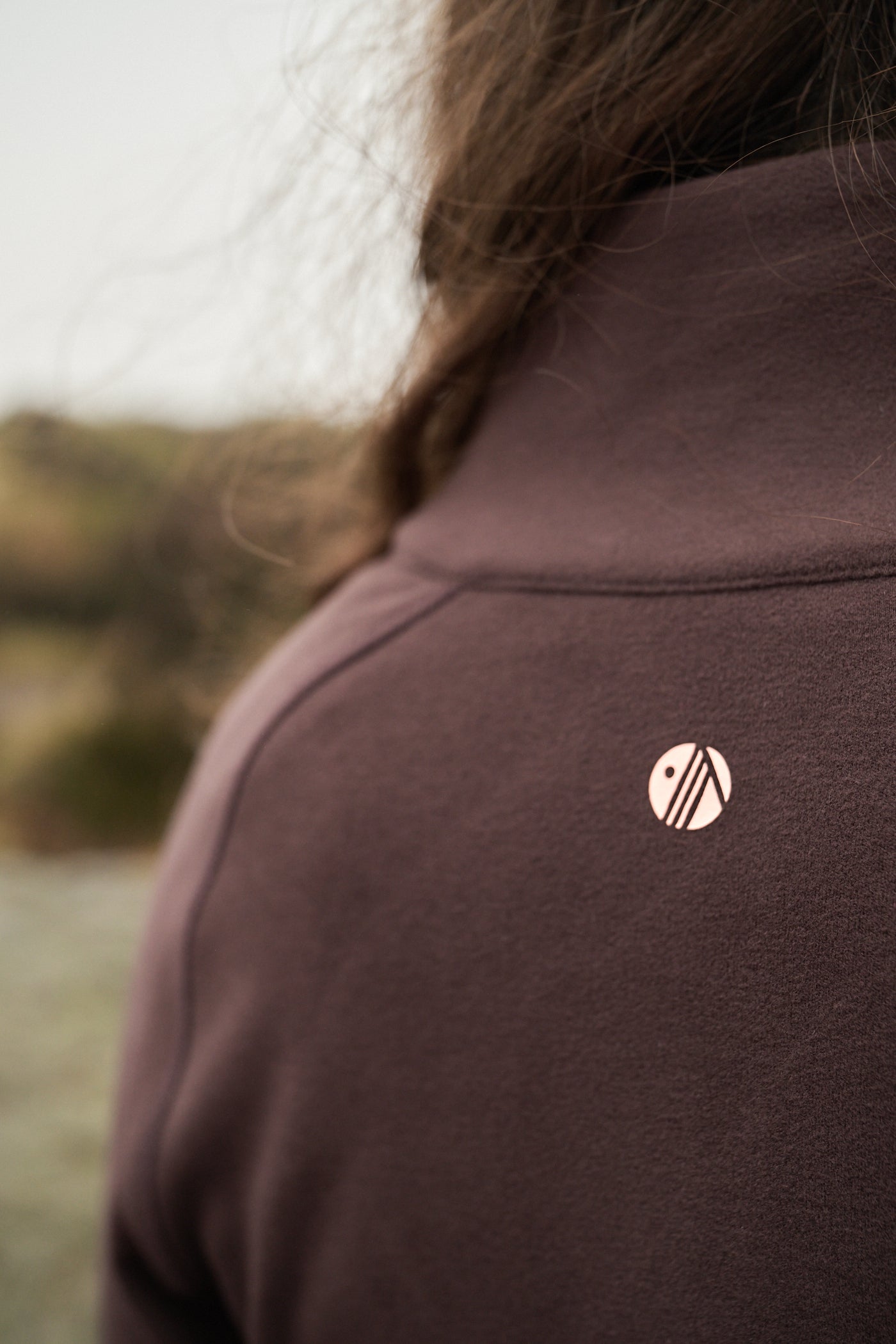 Close-up of a person wearing a brown jacket with a logo on the chest, blurred natural background