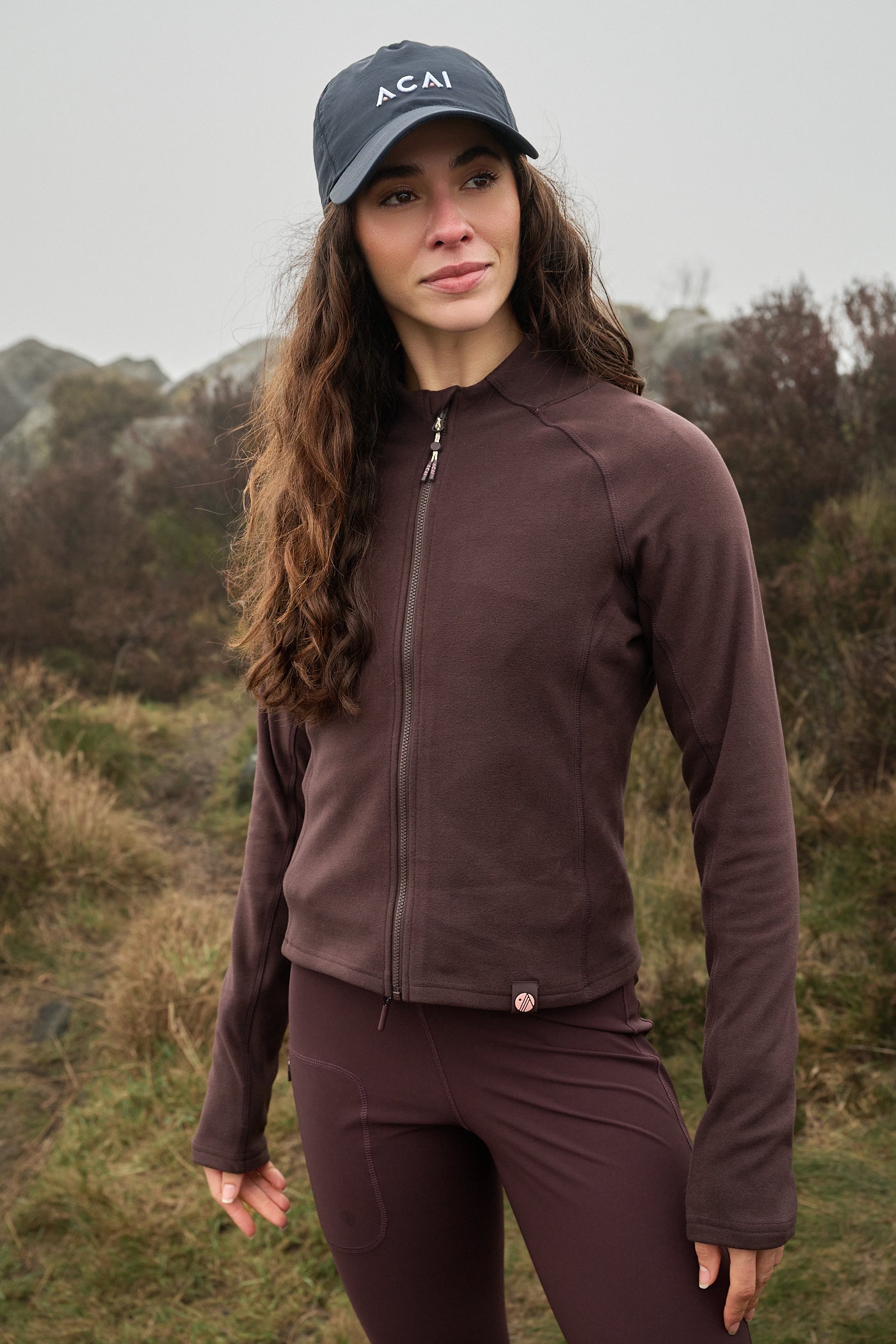 Woman wearing a brown jacket and cap with 'ACAI' branding in a natural setting.