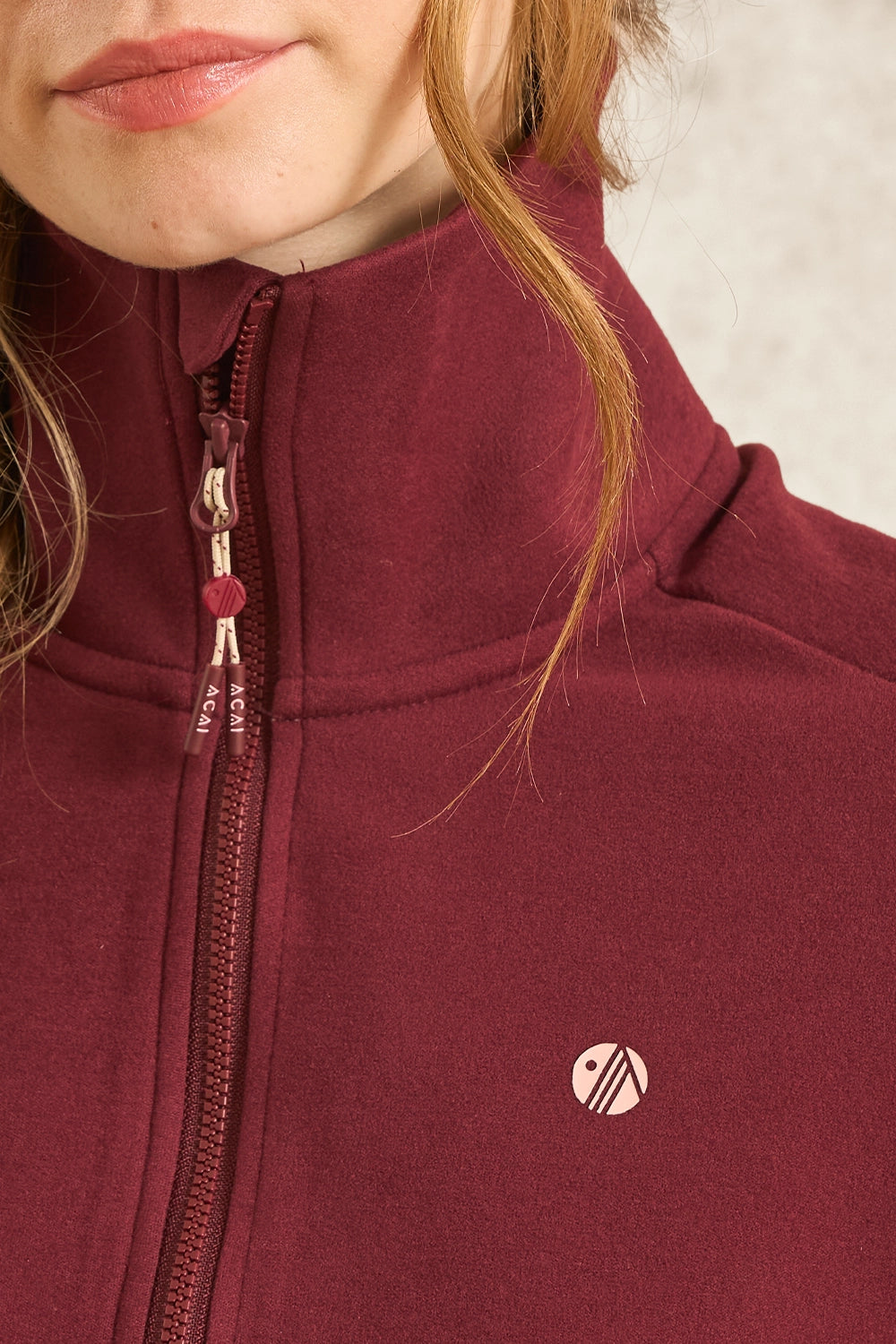 Close-up of a person wearing a burgundy fleece zip-up jacket with a high collar, a matching zipper pull, and a small circular logo on the chest.