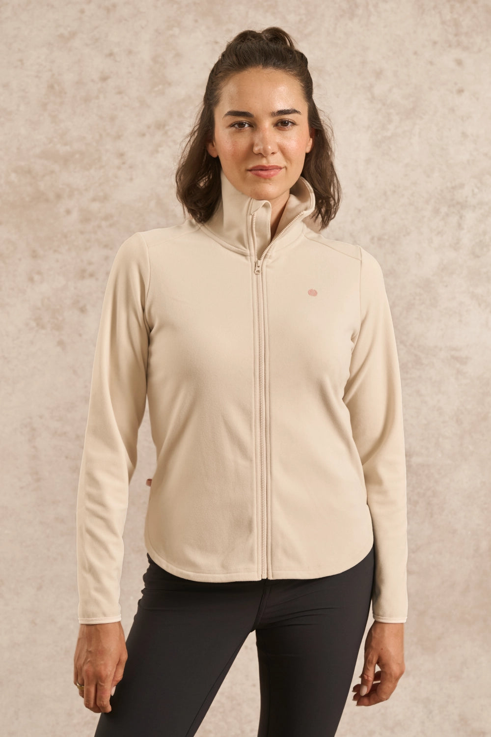 A woman wearing a light beige full-zip fleece jacket with a stand-up collar and black leggings, standing against a textured beige background.
