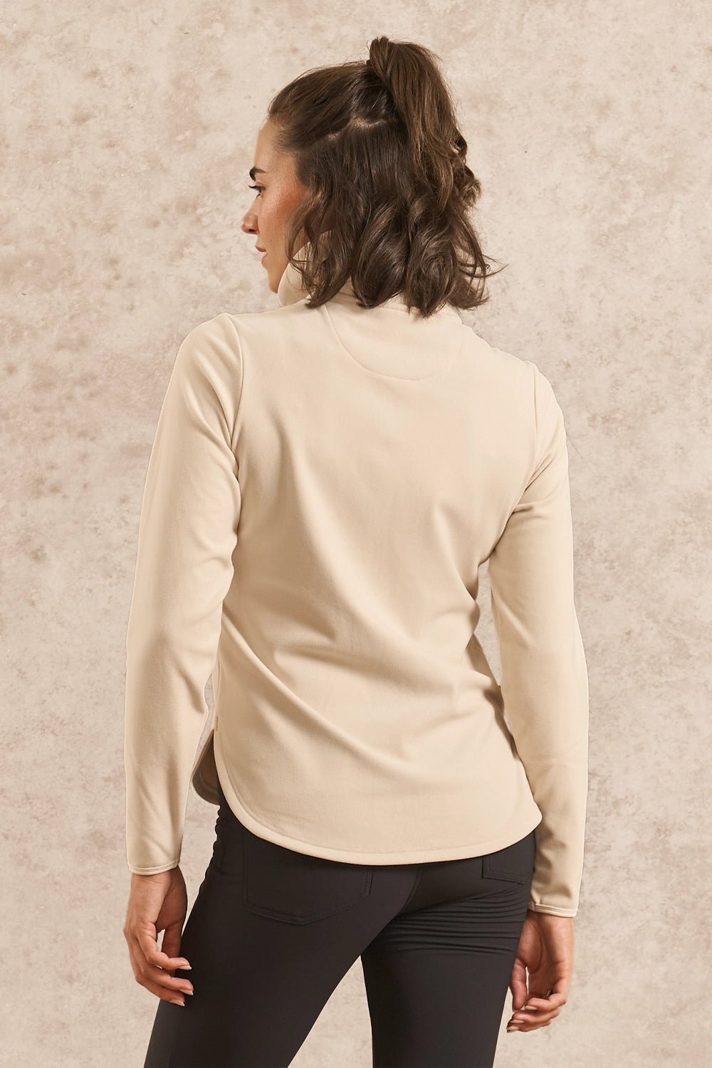 A woman shown from the back wearing a light beige long-sleeve mock neck top and black leggings, standing against a textured beige wall.