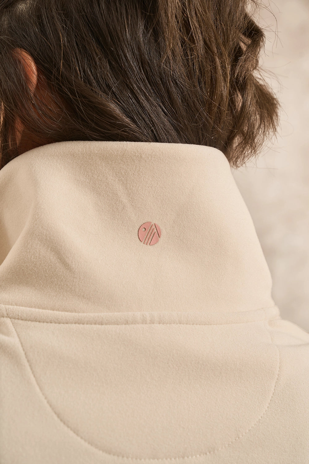Close-up of a person wearing a cream-colored fleece pullover jacket with a high collar and a small pink circular logo on the back.