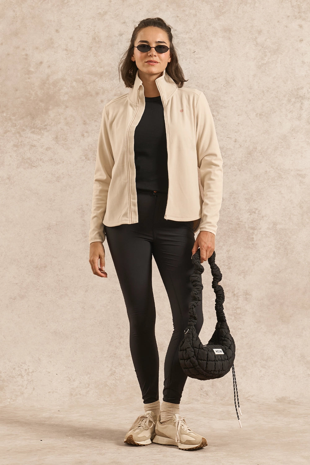 A woman stands against a textured beige studio backdrop wearing a light cream zip-front jacket over a black top and black leggings, with beige sneakers and a black braided shoulder bag.