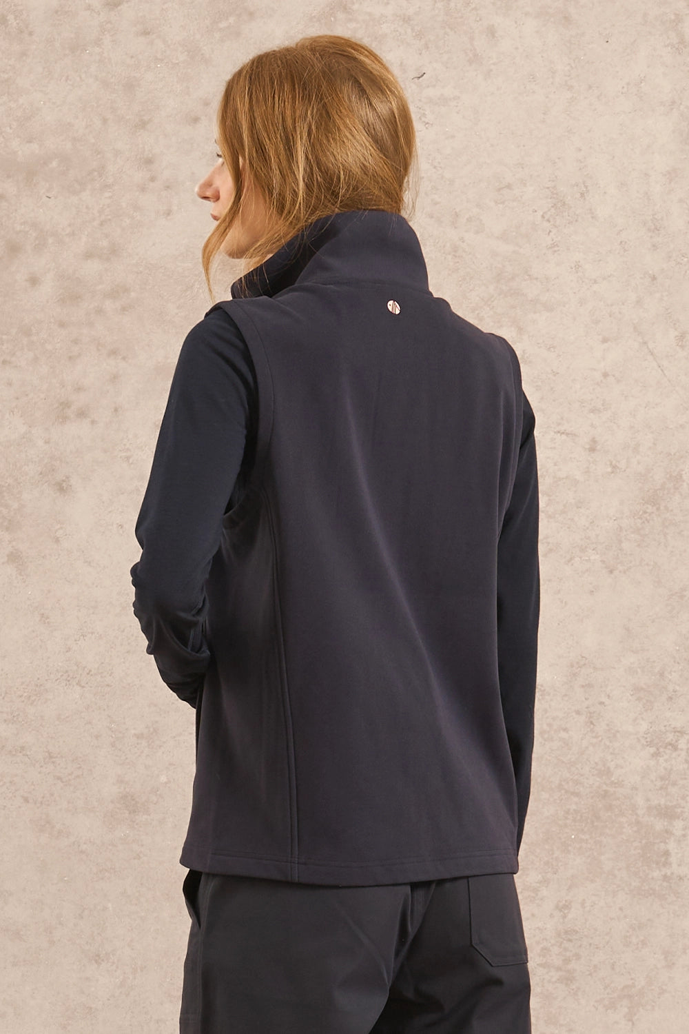 A woman with light brown hair is shown from the back wearing a dark navy fleece vest and matching dark pants, standing against a textured beige wall.