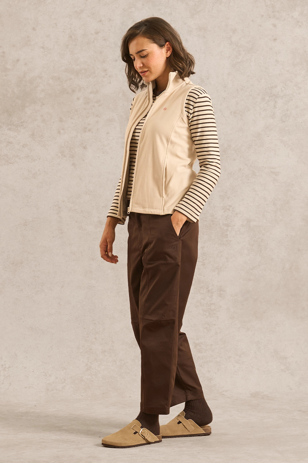 A woman modeling a cream zip-up vest over a black and white striped long-sleeve shirt, paired with dark brown pants and tan slip-on shoes, against a textured light gray studio backdrop.