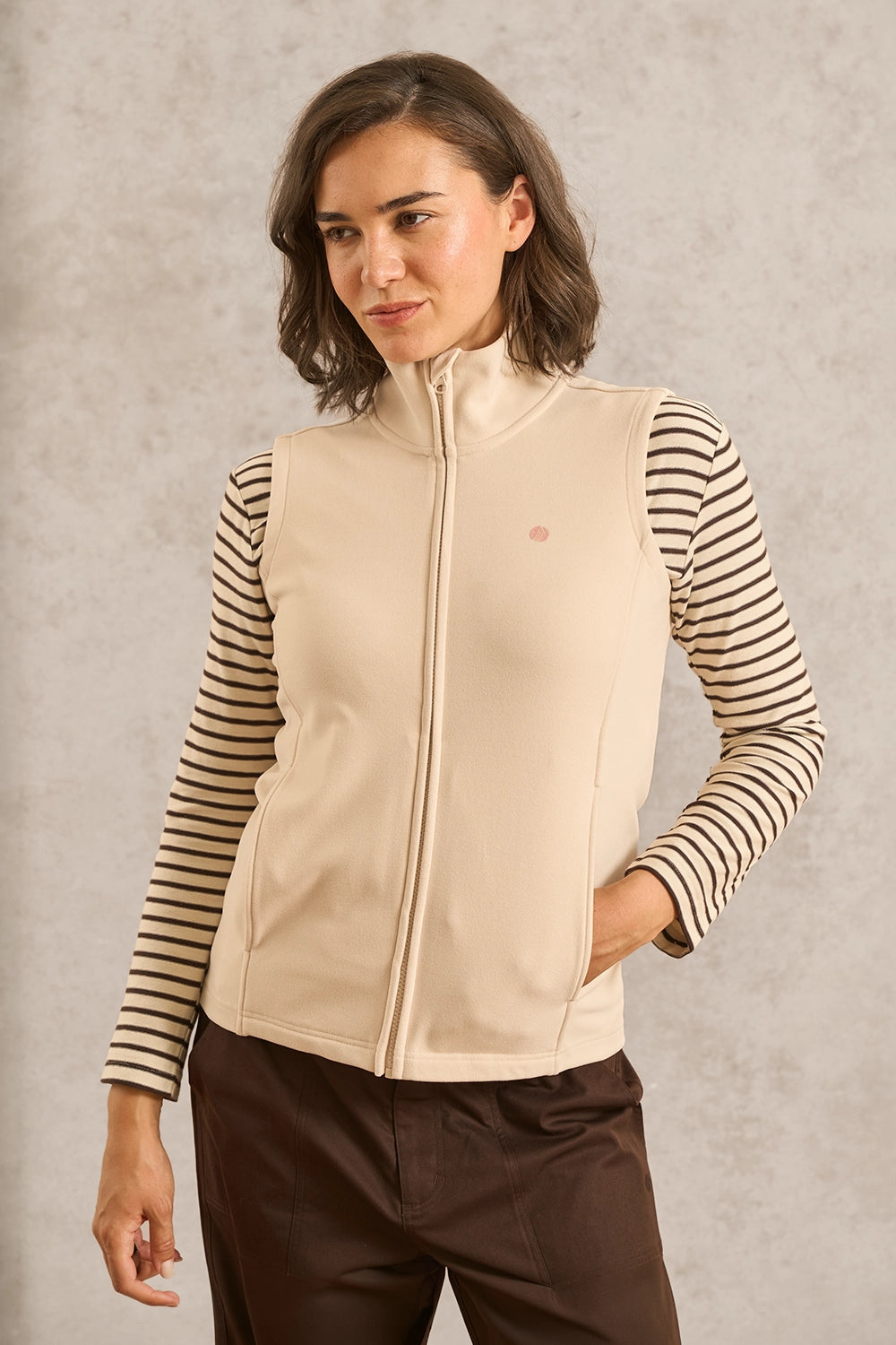 A woman wearing a beige sleeveless zip-front vest over a black and beige striped long-sleeve top, paired with dark brown pants, standing against a textured light gray background.