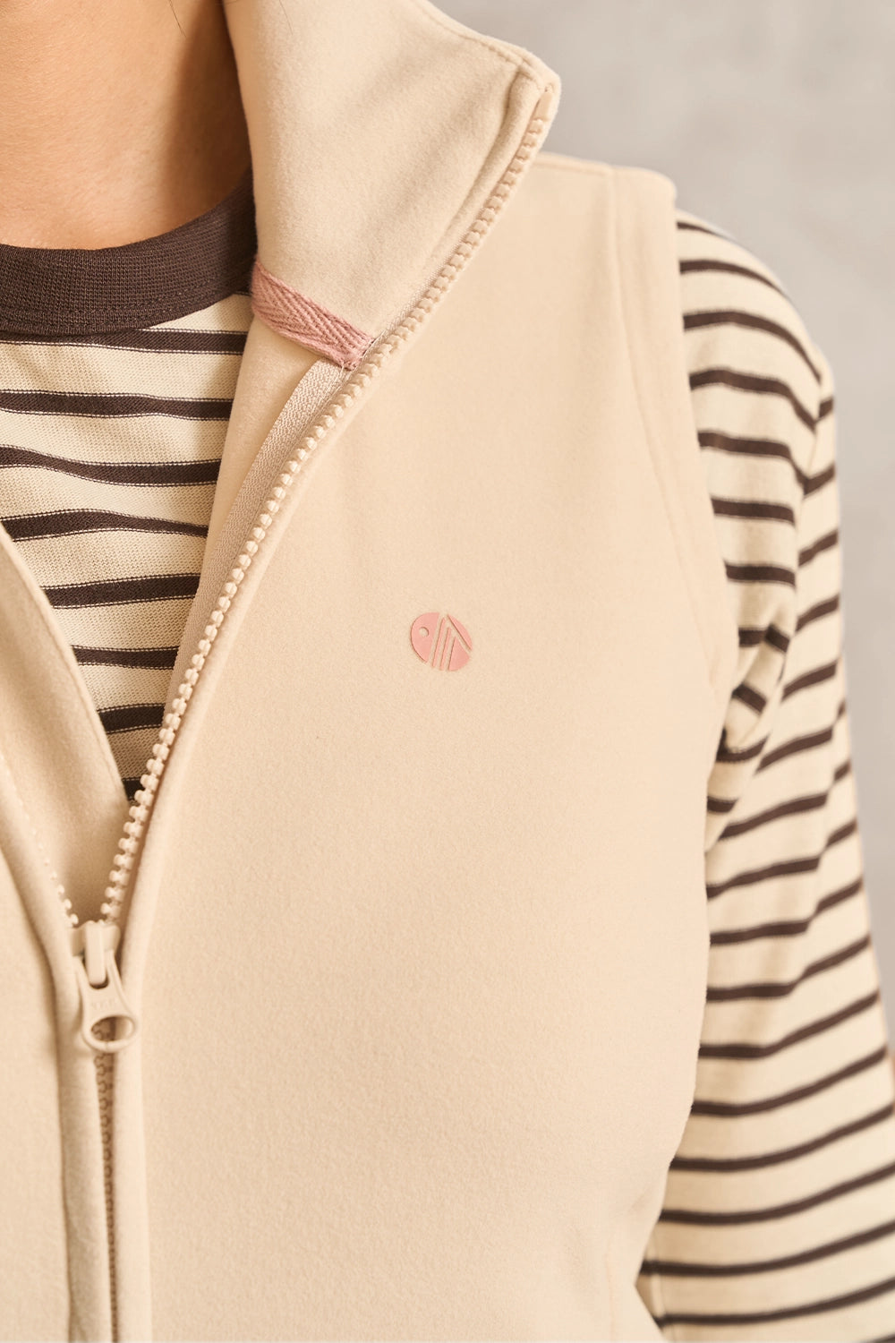 Close-up of a beige zip-front vest worn over a brown and cream striped long-sleeve top. The vest has a small pink embroidered circular logo on the left chest and a light pink zipper trim.