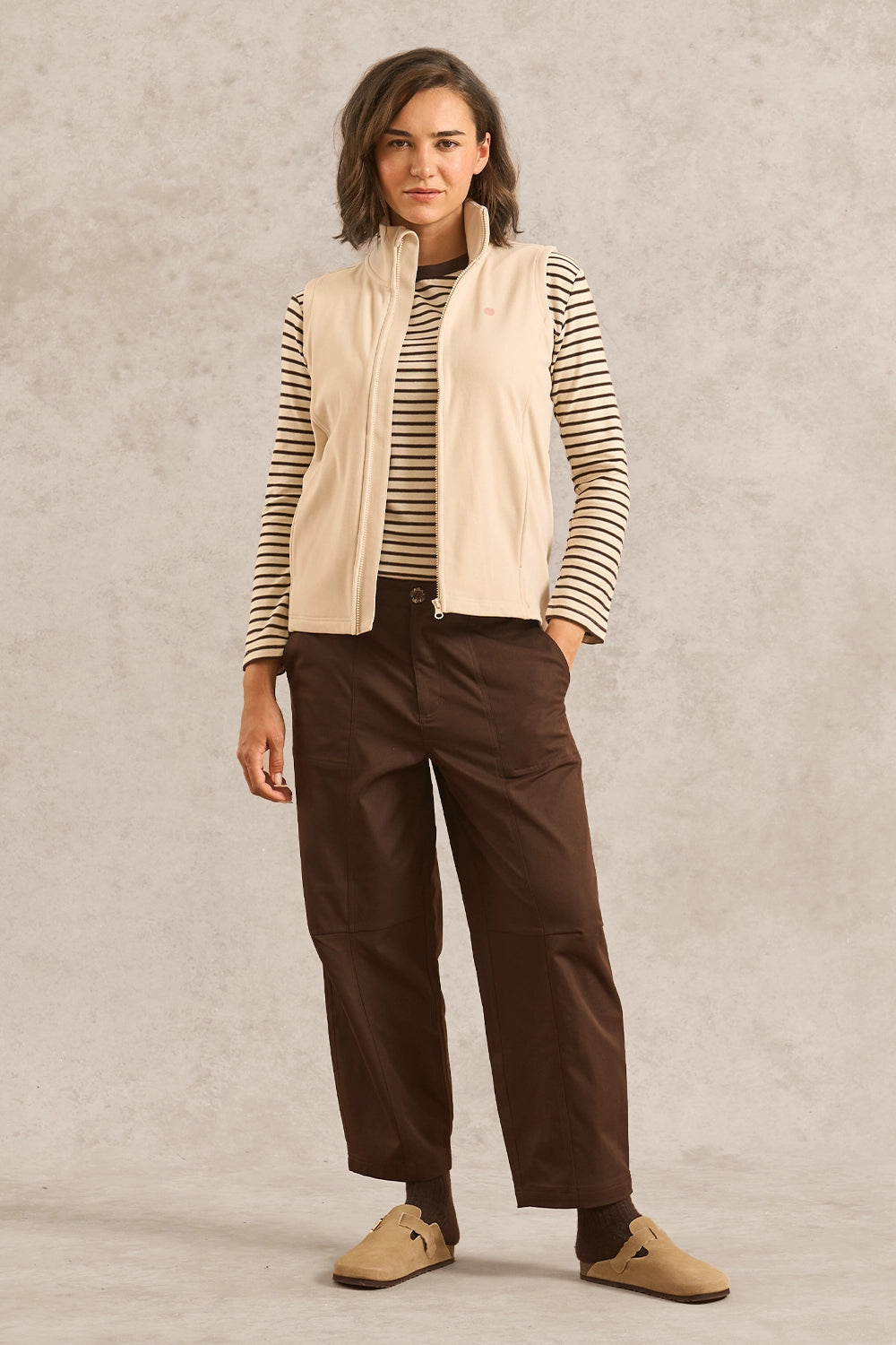 A woman stands against a textured light gray studio backdrop wearing a cream sleeveless zip-front vest over a beige and black striped long-sleeve top, dark brown wide-leg pants, and tan slip-on shoes.
