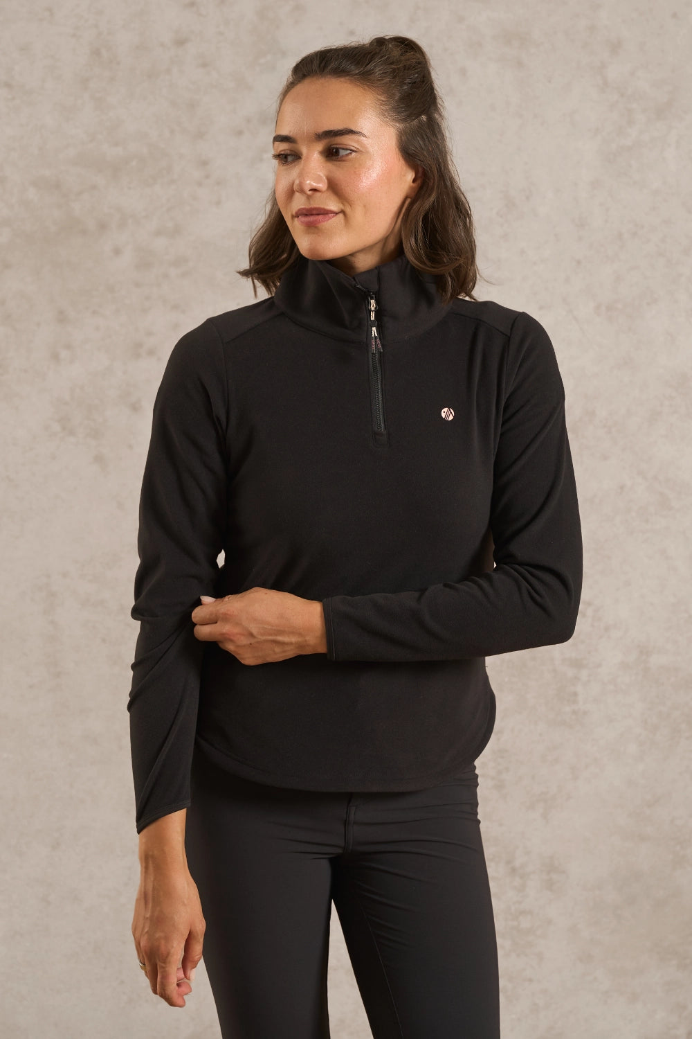 A woman wearing a black long-sleeve quarter-zip athletic pullover with a small circular logo on the left chest, paired with black leggings, standing against a textured light gray wall.