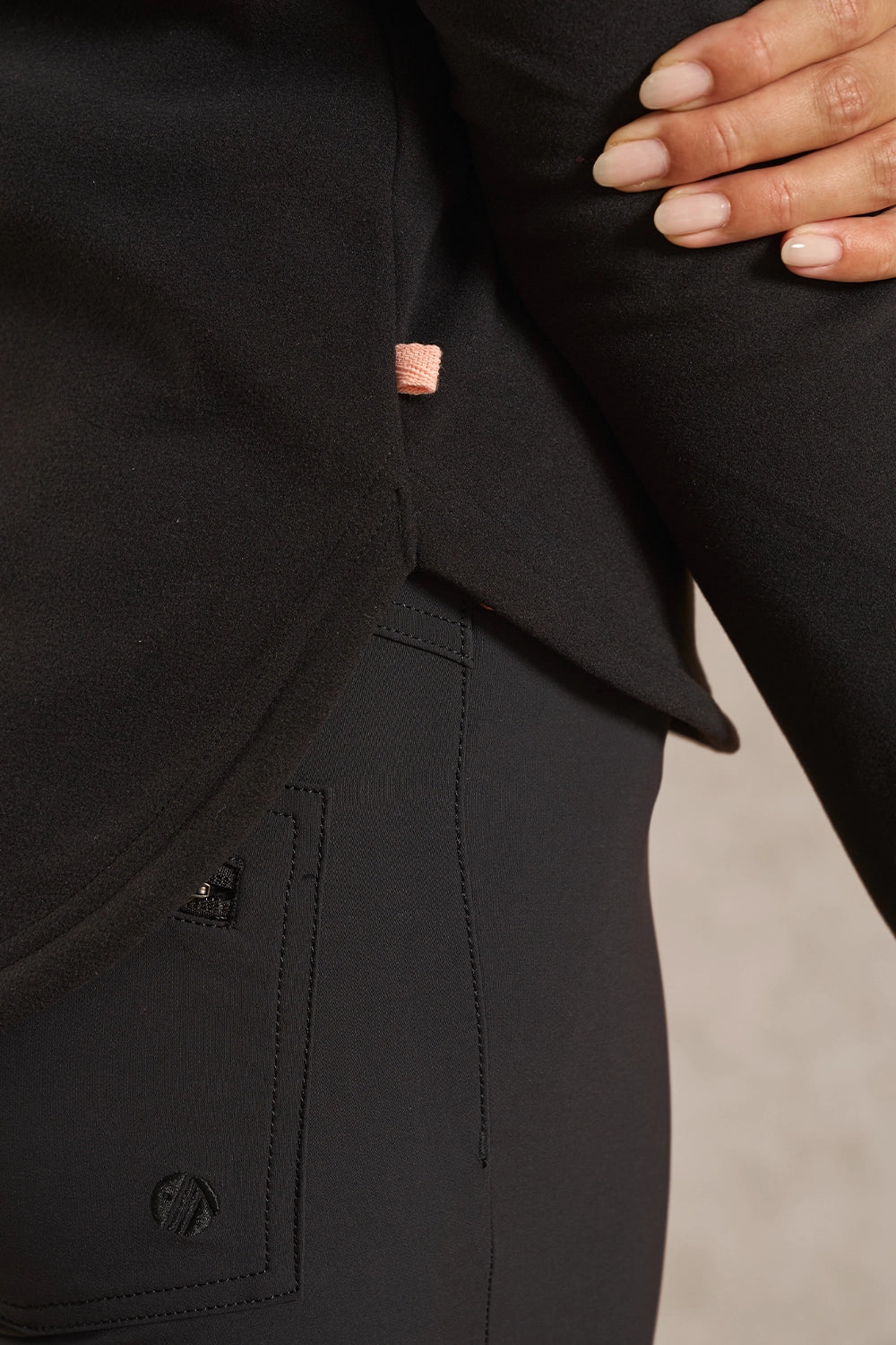 Close-up of a person wearing black athletic apparel, showing a black top with a small peach-pink logo tab and matching black bottoms. A hand with manicured nails rests on the sleeve.