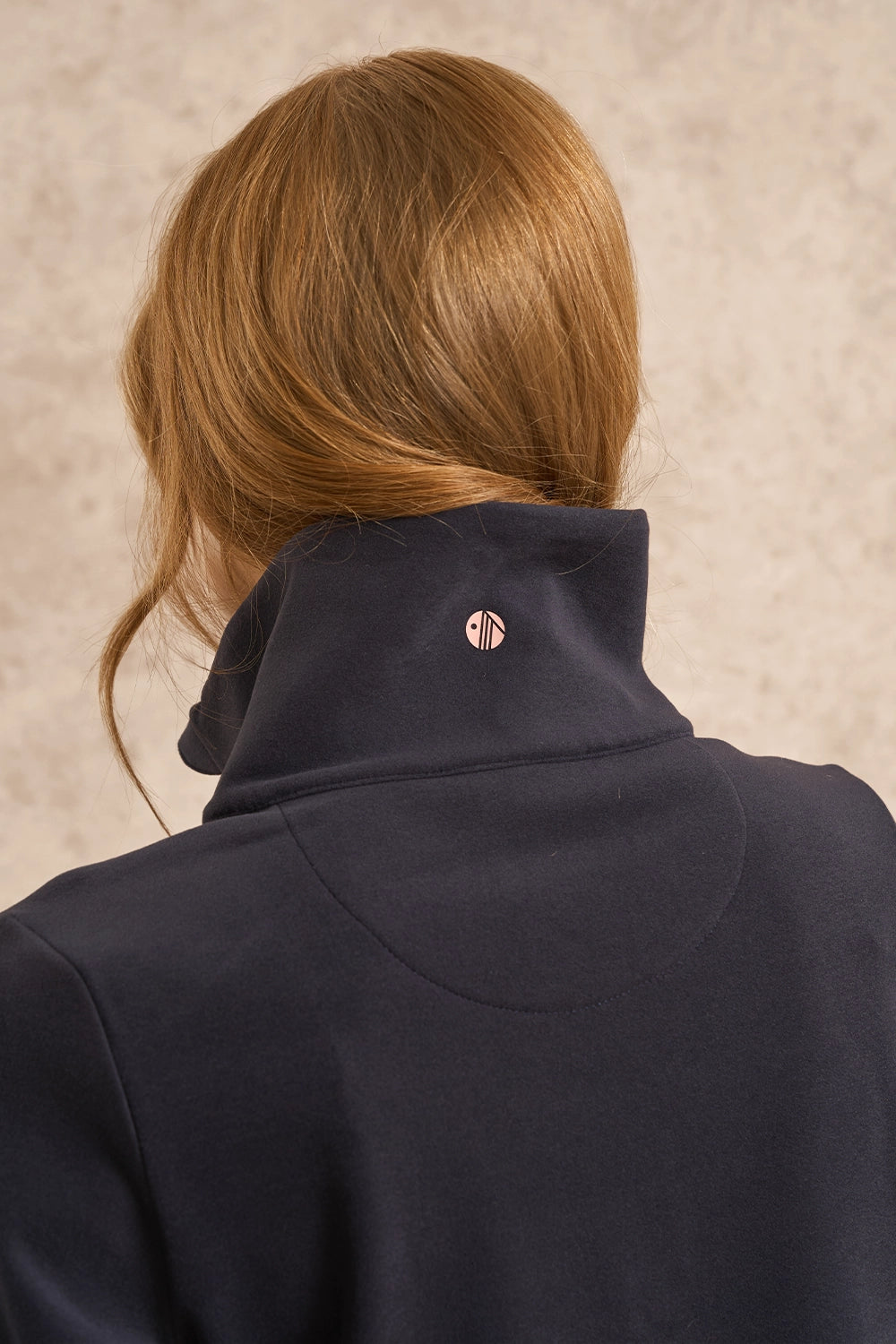 Back view of a person with light brown hair wearing a dark navy high-neck pullover sweater, featuring a small metallic circular logo on the back of the neck, set against a textured beige background.