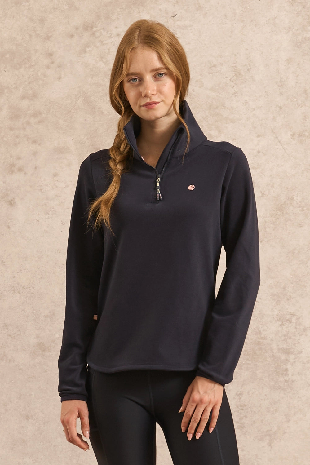 A woman with auburn hair in a braid wearing a dark navy blue quarter-zip pullover sweatshirt and black leggings, standing against a textured beige wall.