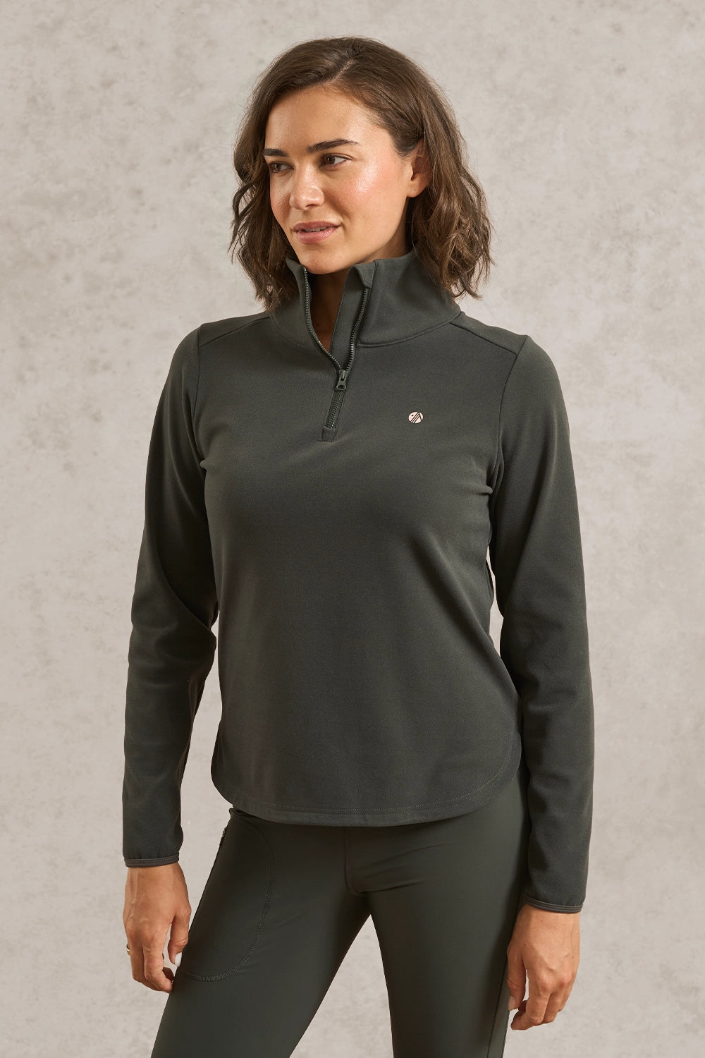 A woman wearing a dark gray long-sleeve quarter-zip athletic pullover and matching dark gray leggings, standing against a textured light gray wall.