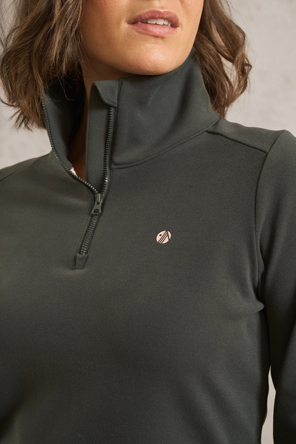 Close-up of a woman wearing a dark olive green quarter-zip pullover with a small circular logo on the left chest, set against a neutral gray background.