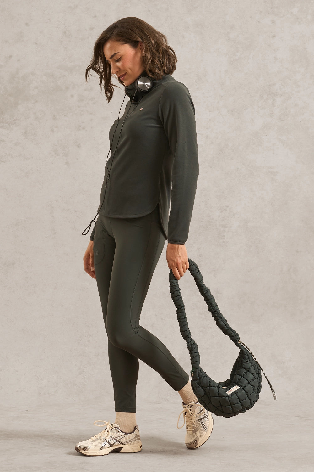 A woman in a dark olive green athletic tracksuit (long-sleeve top and leggings) is shown from the side, holding a dark green braided shoulder bag. She is wearing beige sneakers against a textured light gray studio background.
