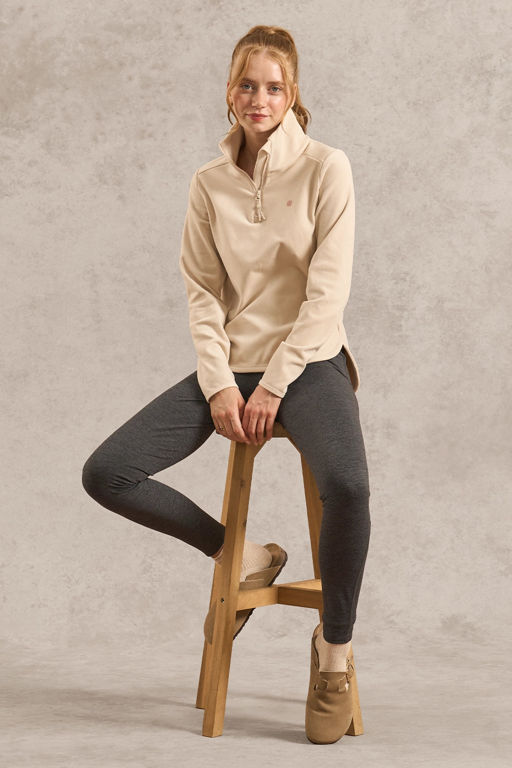 A woman sits on a wooden stool wearing a beige quarter-zip fleece pullover, dark gray leggings, and tan lace-up boots against a textured gray studio backdrop.