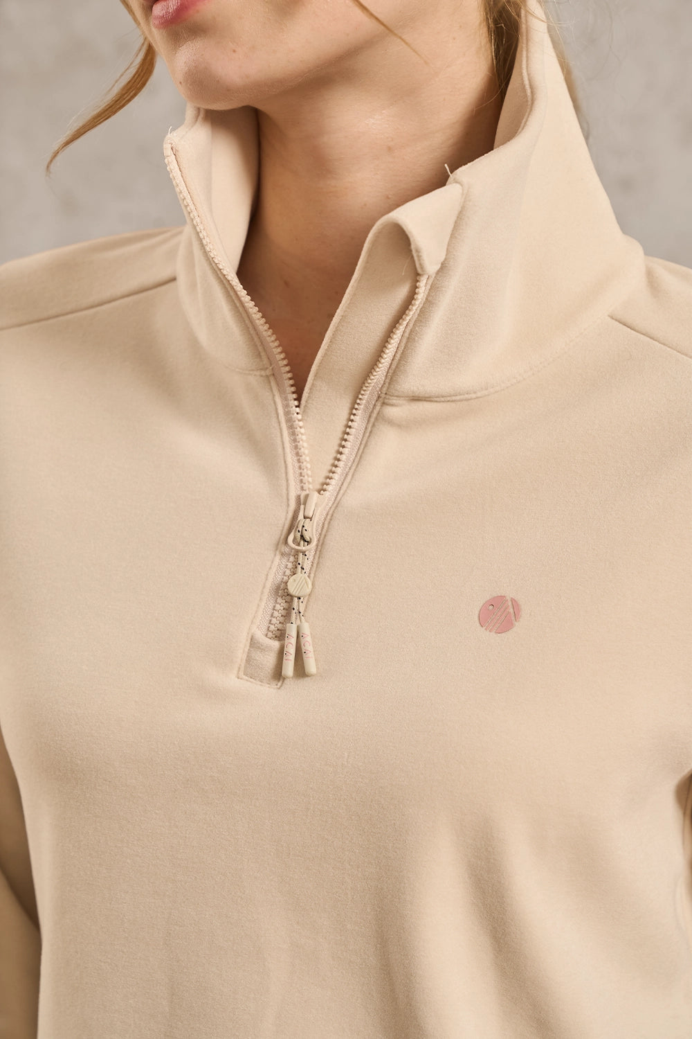 Close-up of a model wearing a beige quarter-zip pullover sweater with a small pink circular logo on the left chest.