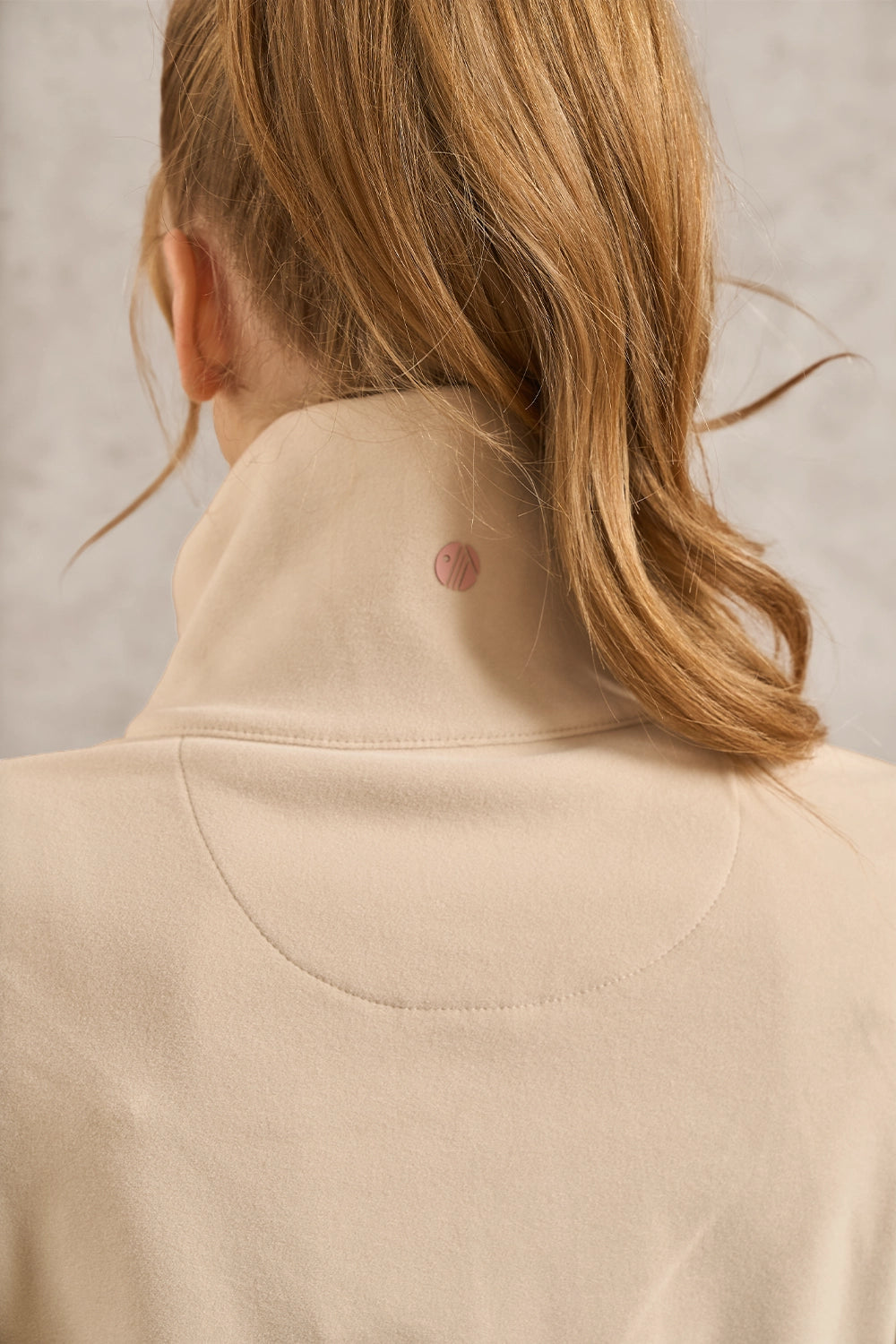 Close-up back view of a woman wearing a beige high-neck top, showing the neck and hair against a light textured background.