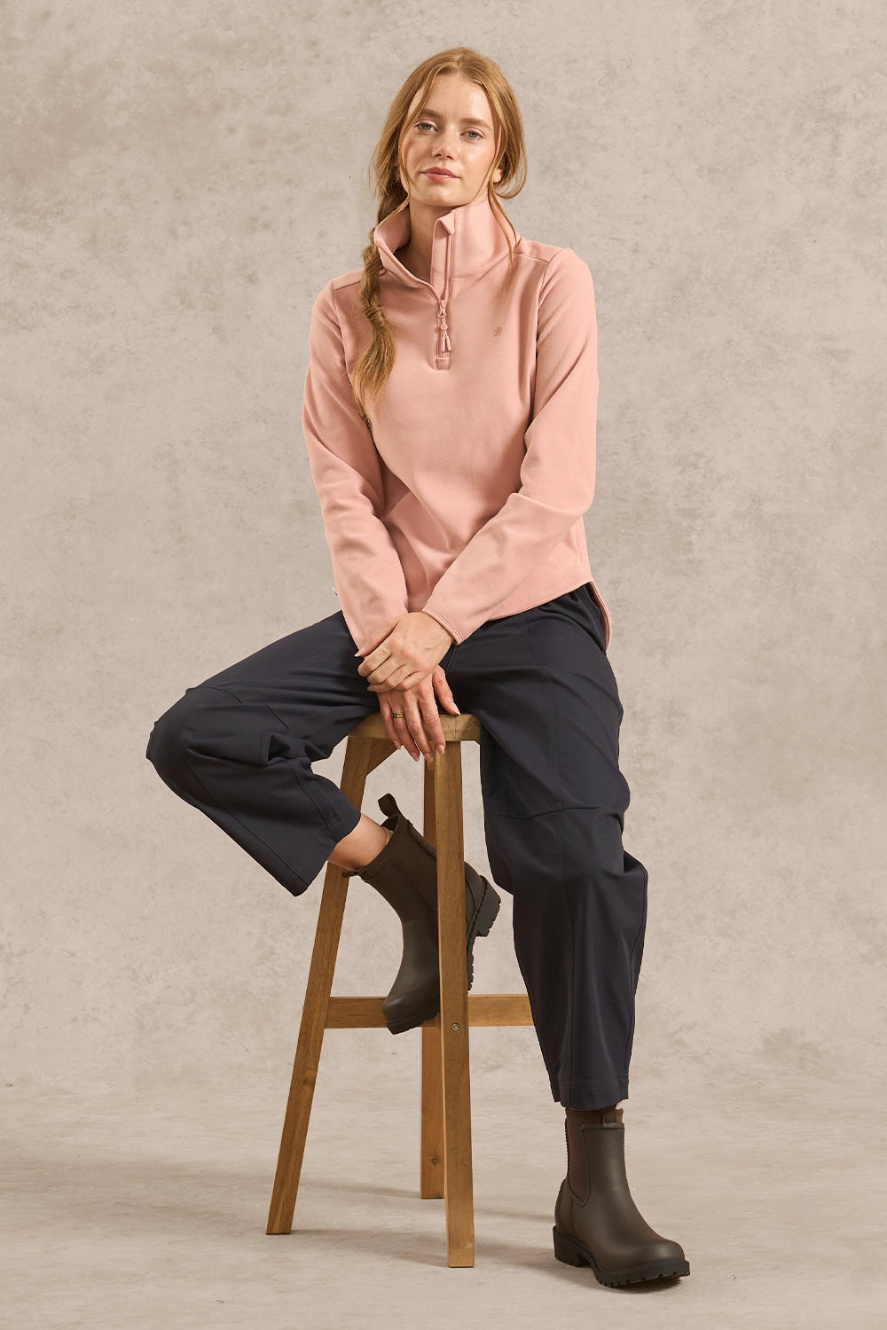 A woman sits on a wooden stool wearing a dusty pink quarter-zip fleece pullover, dark navy cropped trousers, and dark brown boots against a textured light gray studio backdrop.
