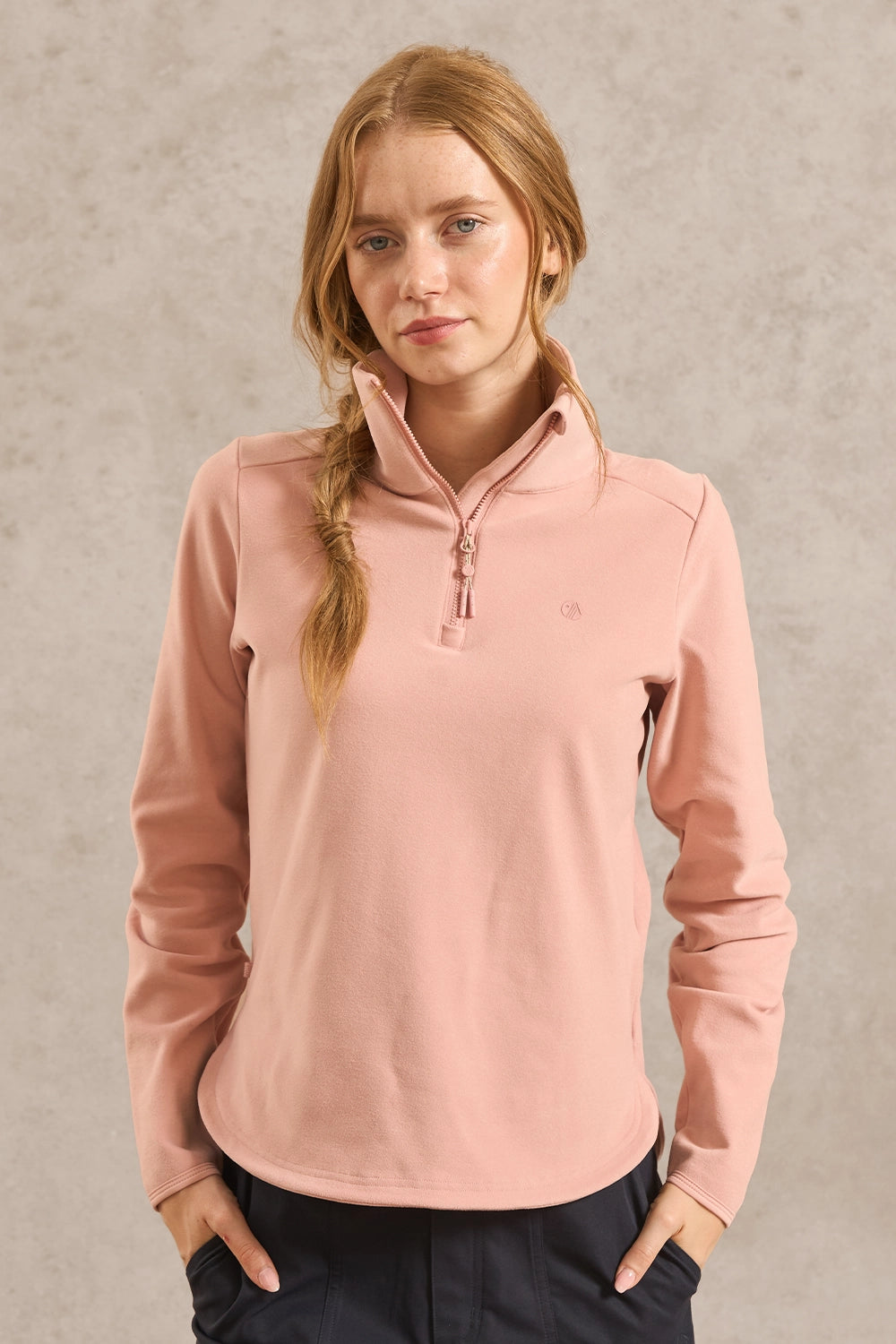 A woman with blonde hair in a braid wearing a dusty pink quarter-zip pullover sweater and dark navy pants, standing against a textured gray wall.