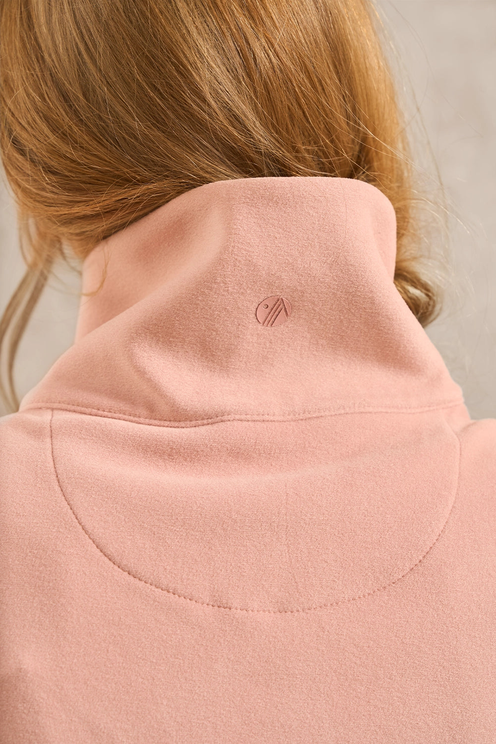 Close-up back view of a person wearing a dusty pink fleece turtleneck sweater, showing the high collar and a small tonal embroidered logo on the back.