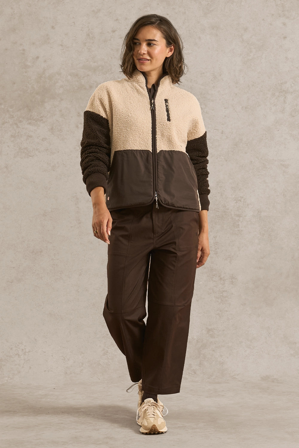 A woman modeling a two-tone fleece zip-up jacket and matching dark brown pants against a textured gray studio backdrop.