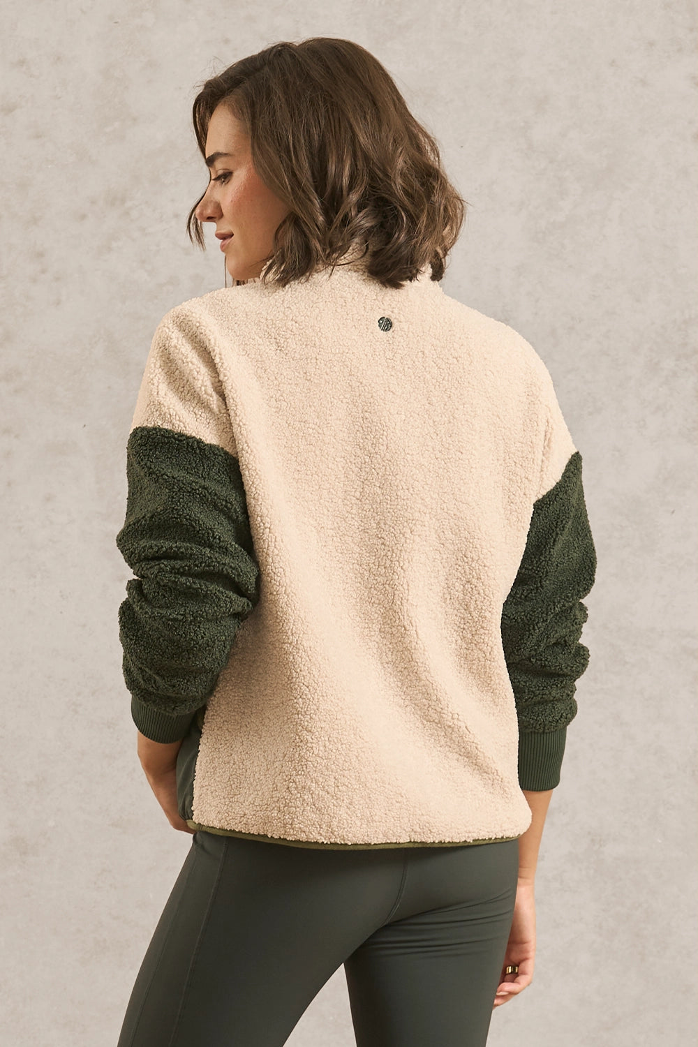 A woman shown from the back wearing a color-block fleece pullover with a cream-colored body and dark green sleeves, paired with dark green leggings, against a textured light gray wall.
