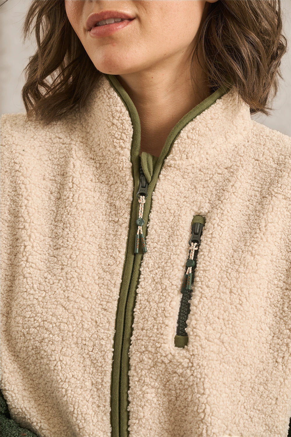 Close-up of a person wearing a cream sherpa fleece zip jacket with olive green trim and zipper pulls.