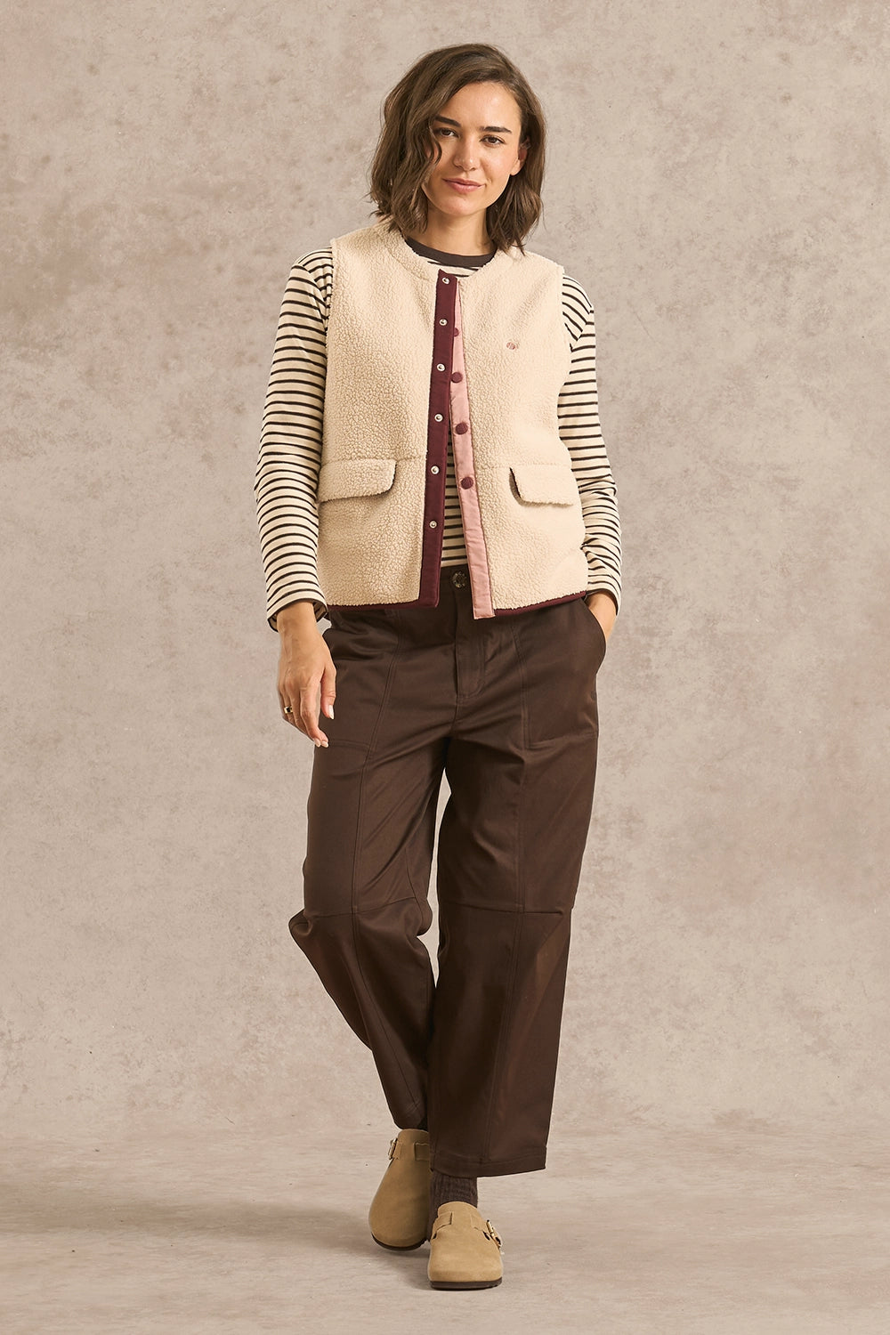 A woman modeling a cream sherpa vest over a striped long-sleeve shirt and dark brown pants, standing against a textured beige studio backdrop.